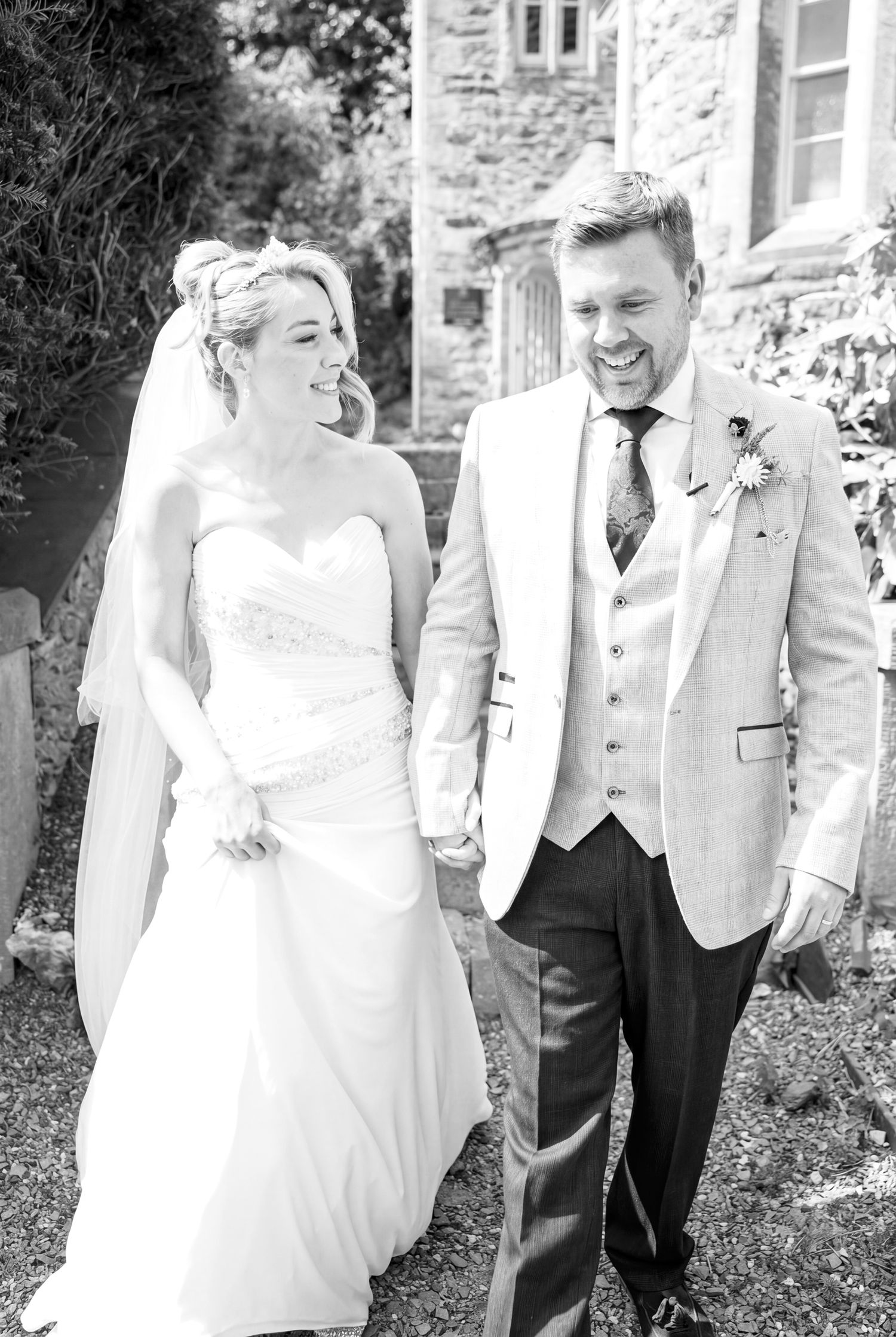Black and white photo of a smiling bride and groom holding hands walking outside a stone building on their wedding day.