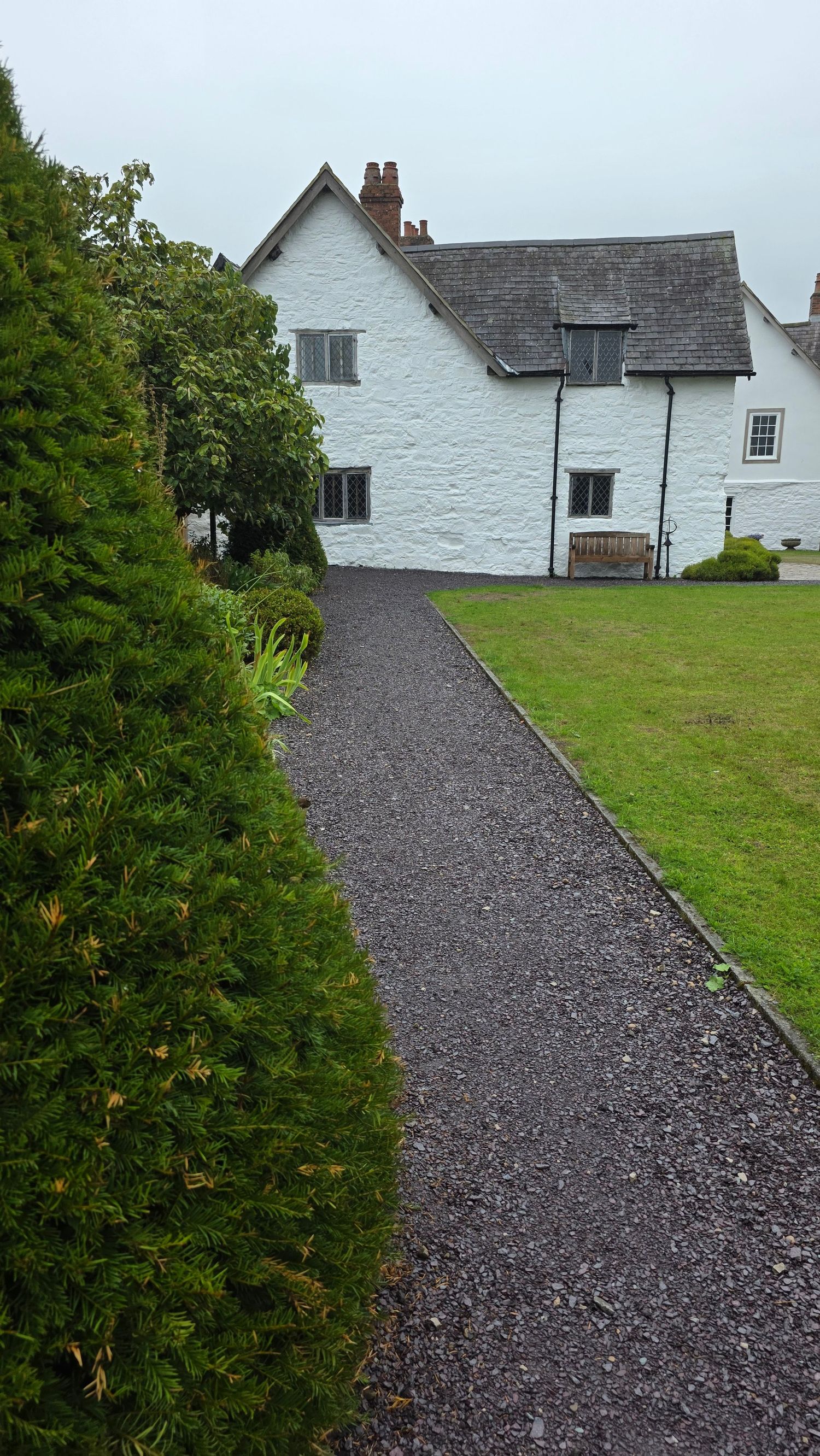 Gravel path leading to a whitewashed country cottage with slate roof, surrounded by manicured lawn and evergreen shrubs.