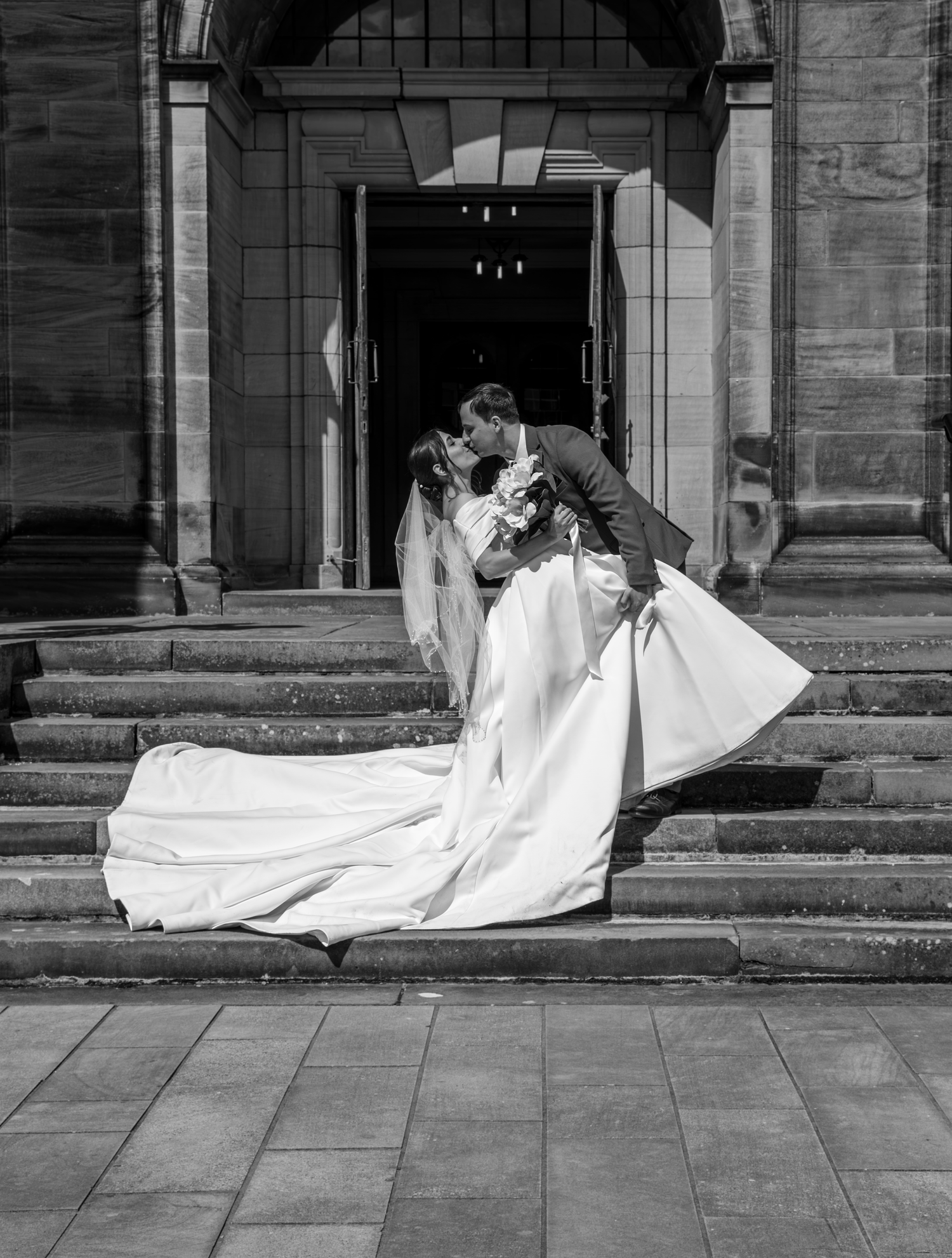 Black and white photo of a bride and groom sharing a romantic dip kiss on stone steps outside a grand church.