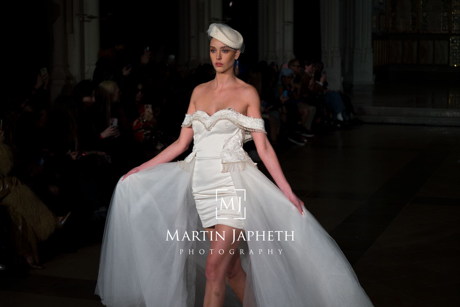 Model walks runway in dramatic white wedding gown with detachable overskirt during fashion show.