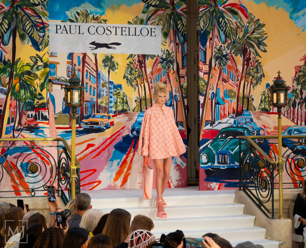 Colorful fashion runway show with models walking past vibrant tropical mural backdrop featuring palm trees and bright designs.