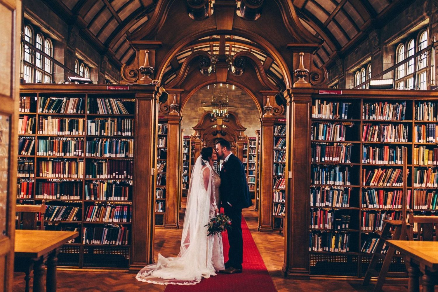 Bride and groom share a romantic kiss in an elegant library with floor-to-ceiling bookshelves and a red carpet aisle. Bangor University