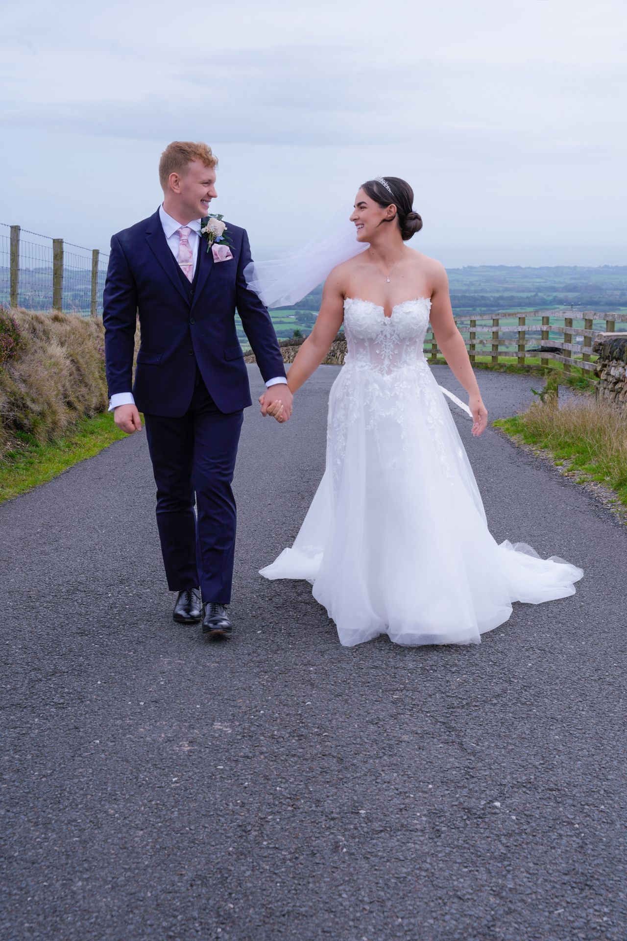 Bride and groom walking hand in hand on a country road, smiling at each other, bride in white gown, groom in navy suit.