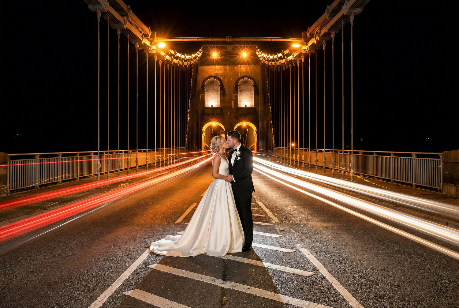 A couple in wedding attire stands on an illuminated Menai Suspension Bridge at night with light trails from passing cars.