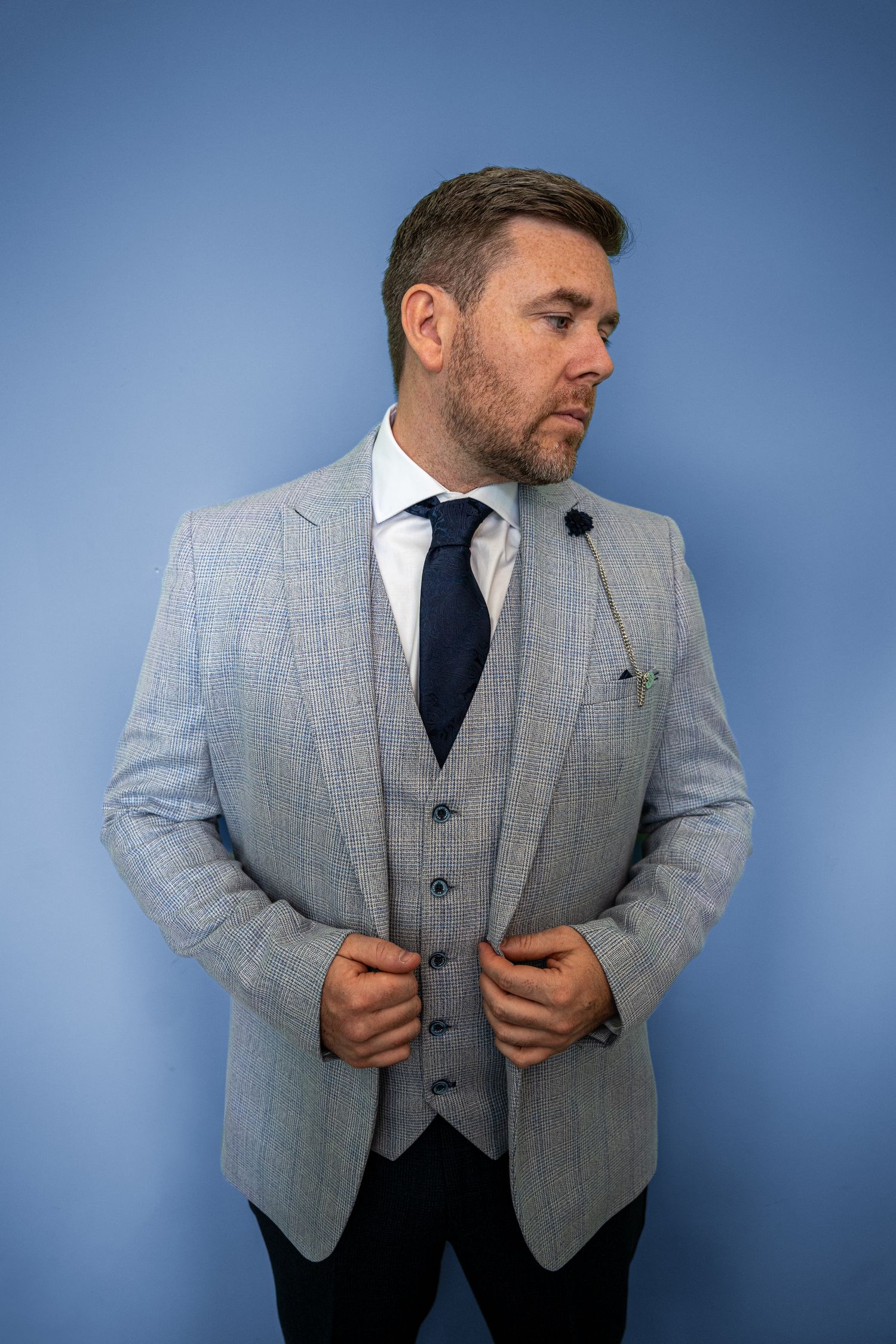 Liam in his light gray checked suit with navy tie and lapel pin poses against blue background.