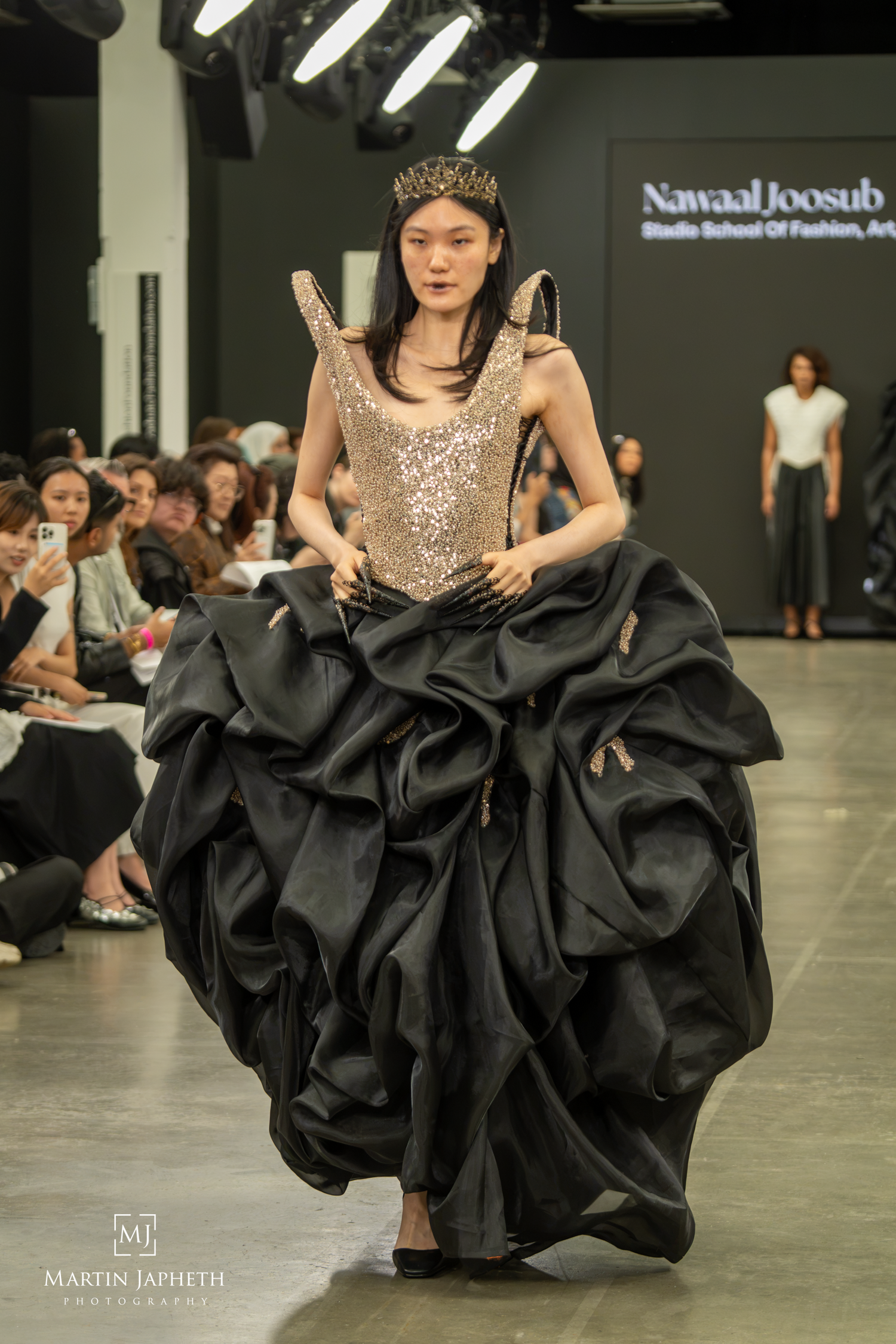 Model walks runway in gold sequin bodice and dramatic black ruffled ball gown skirt with crown at fashion show. Martin Japheth Photography