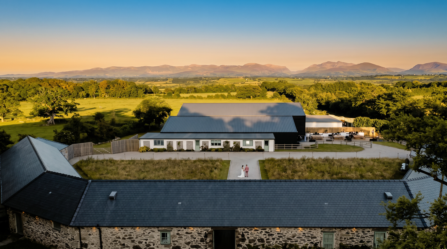 Aerial view of a modern countryside venue with mountain backdrop at golden hour, surrounded by lush green fields.