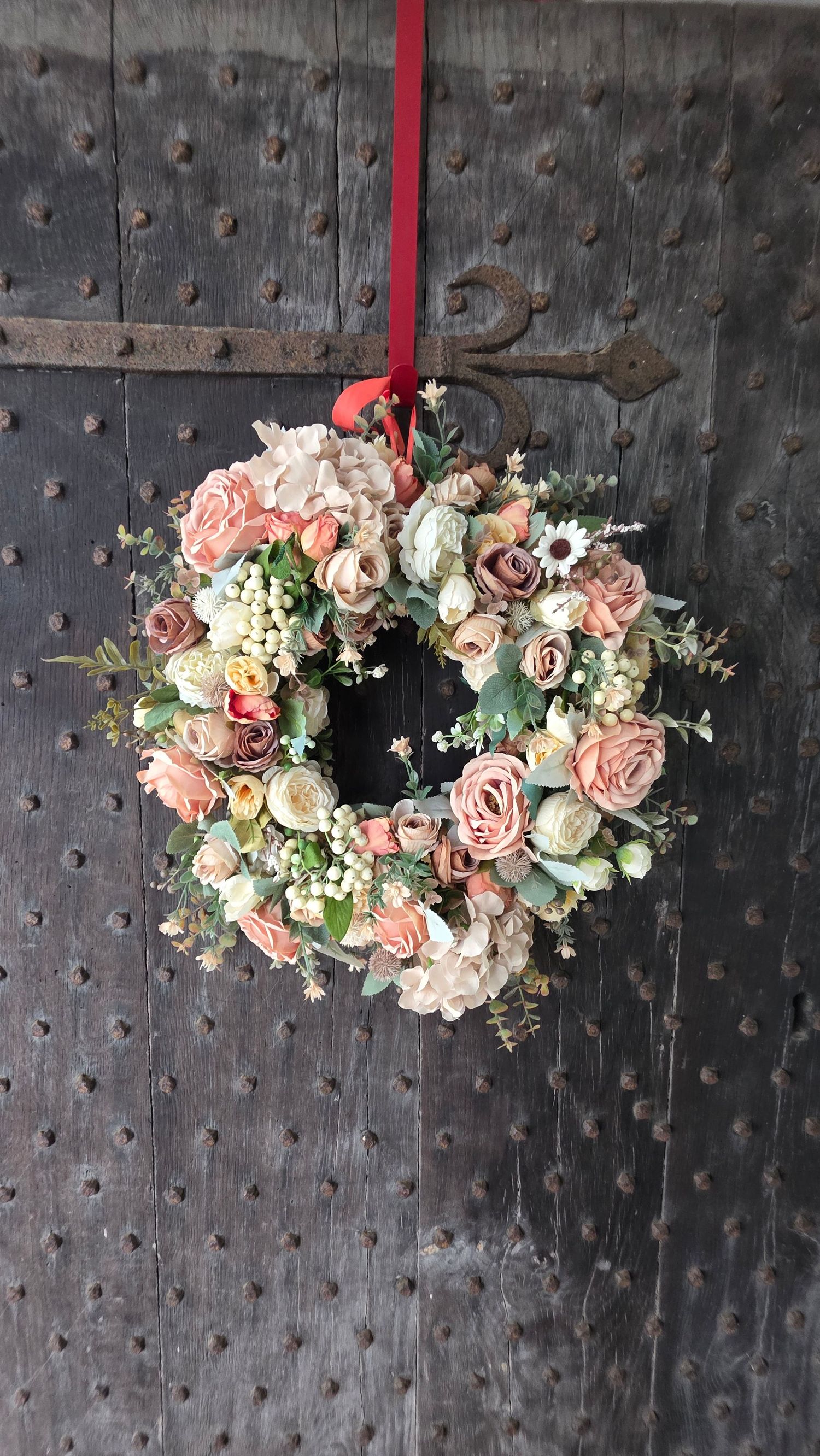 Elegant floral wreath with peach roses and white blooms hanging on a rustic wooden door with a red ribbon.