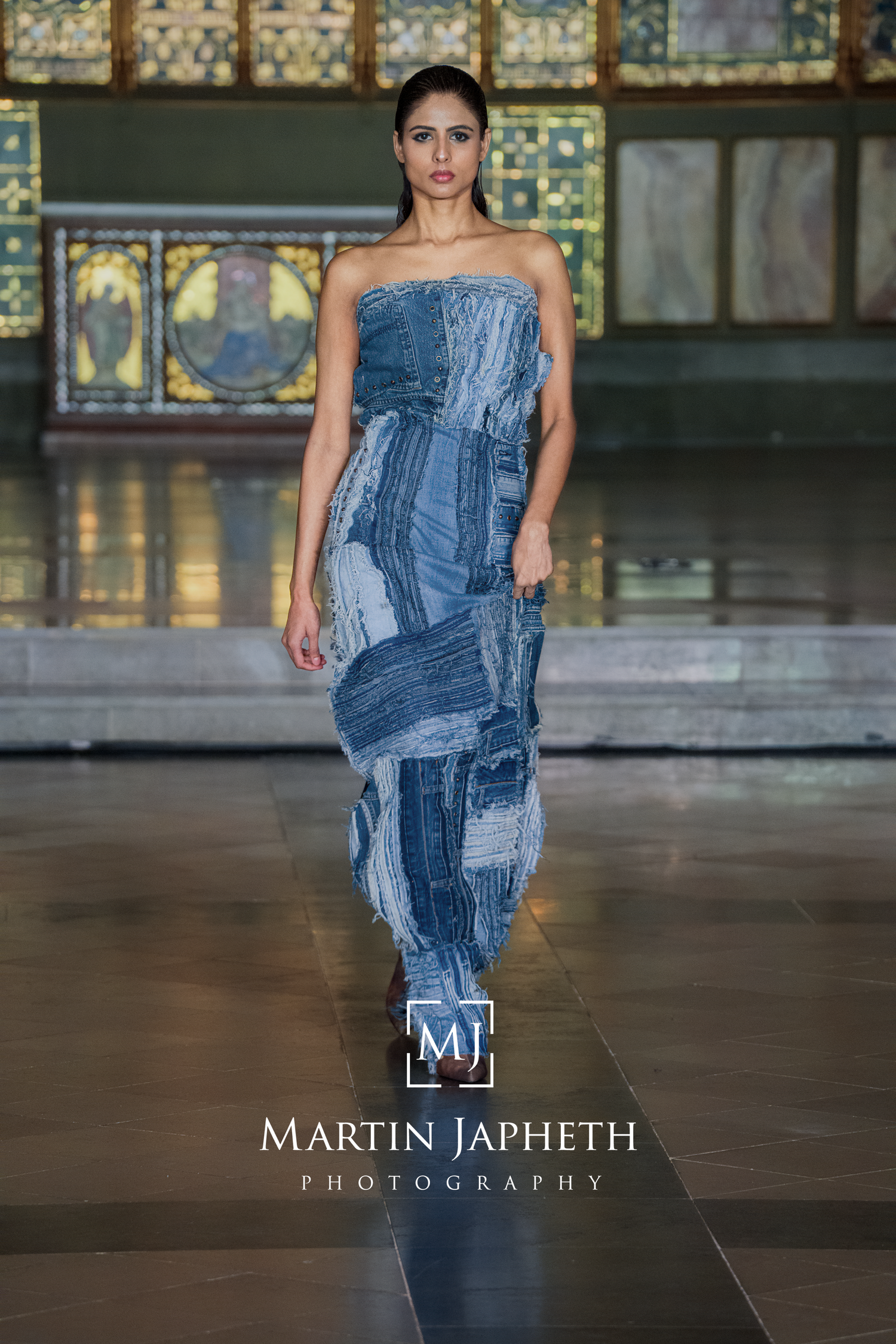 Model showcases strapless patchwork denim evening gown on fashion runway with stained glass windows in background.