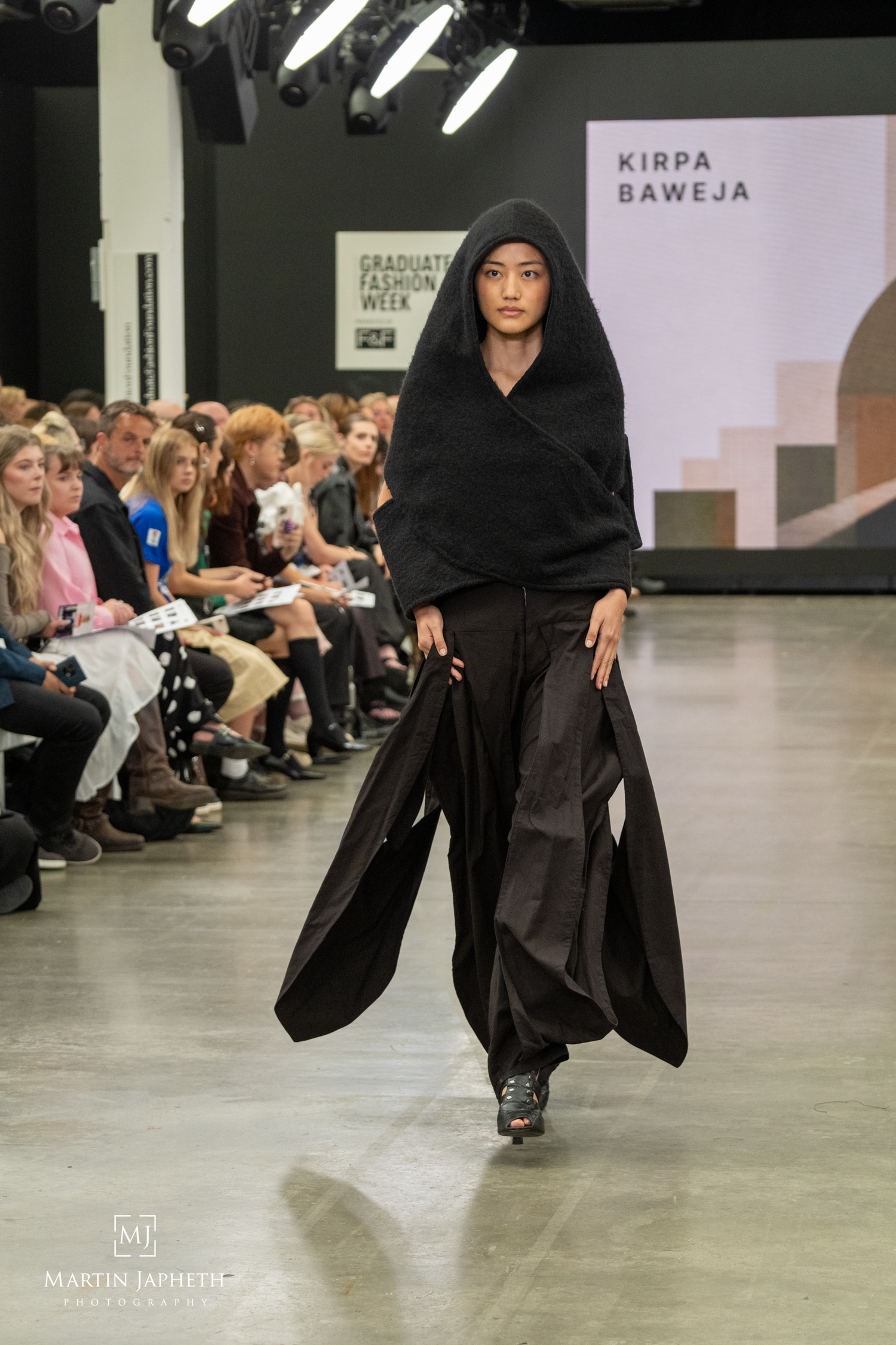 Model walks runway in all-black avant-garde ensemble at Kirpa Baweja Graduate Fashion Week show. Martin Japheth Photography