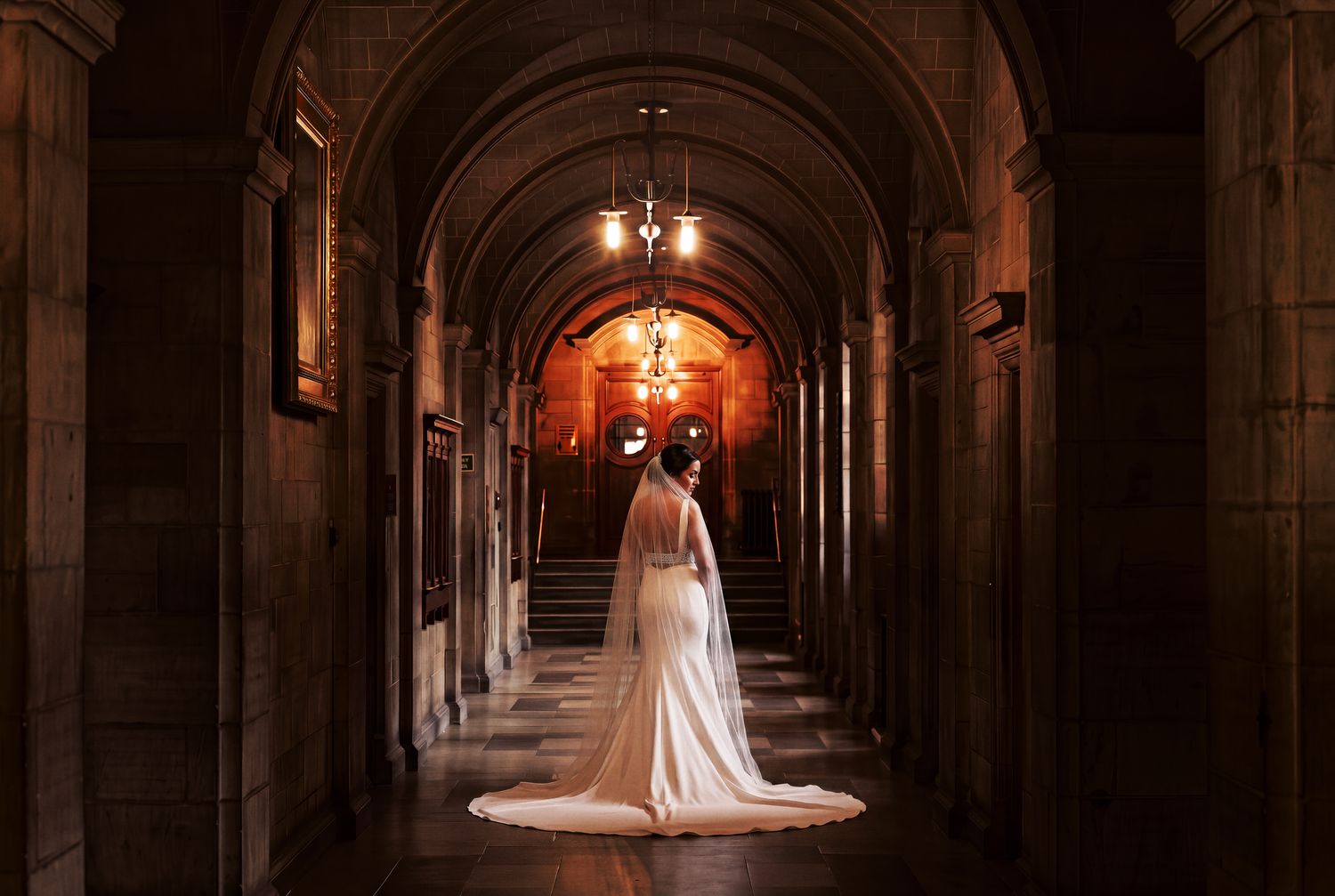 Bangor University Wedding Bride in elegant white gown and veil stands in ornate stone hallway | Martin Japheth Photography