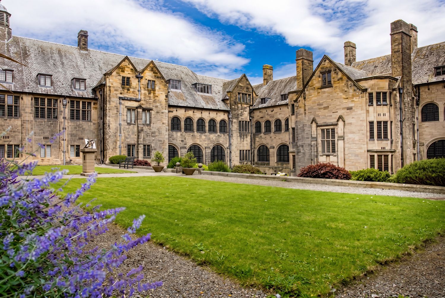 Historic stone manor house with Gothic architecture surrounds a lush green courtyard with purple flowers under a blue sky.