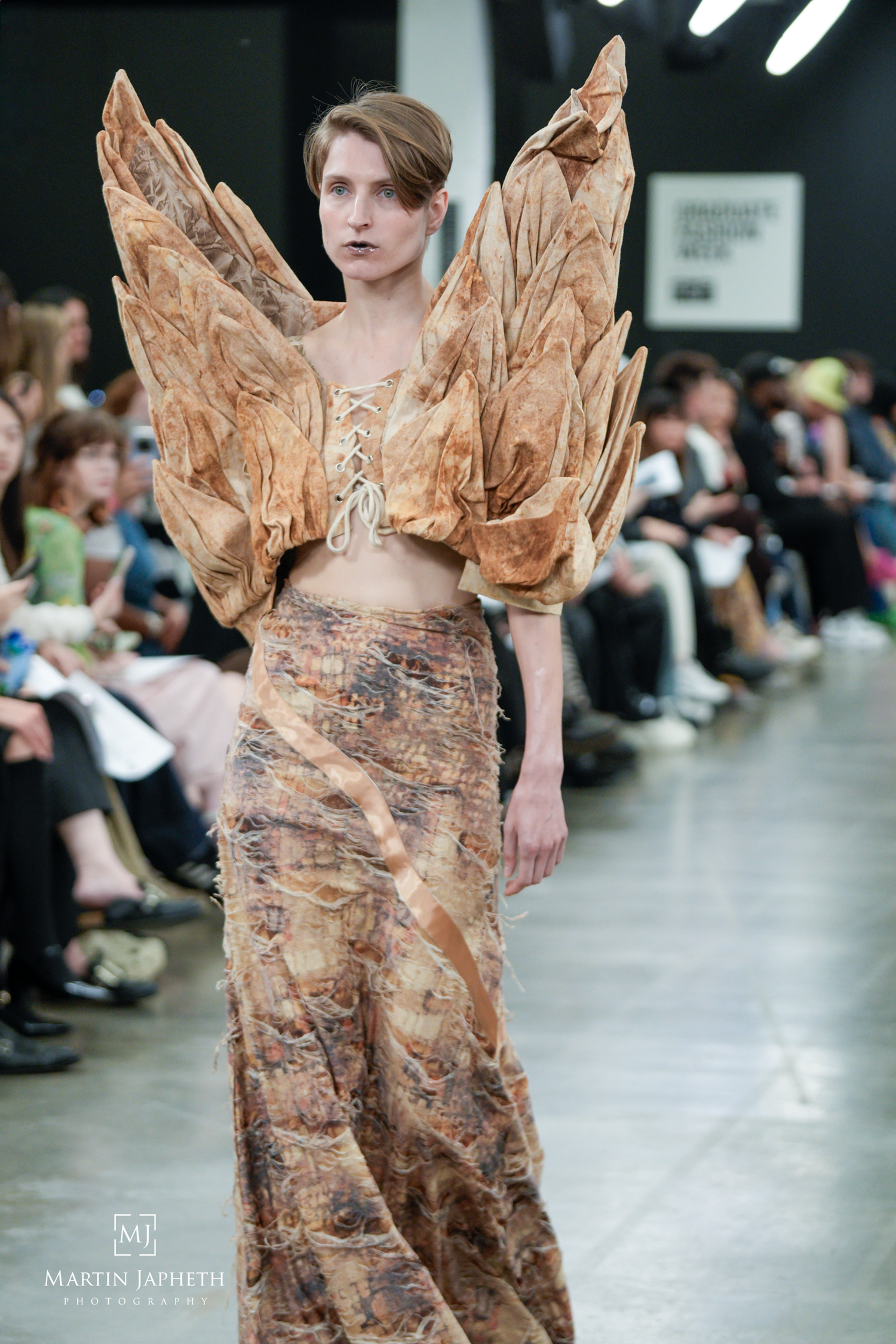 Model walks runway in avant-garde brown leaf-textured winged top and flowing maxi skirt at fashion show. Martin Japheth Photography