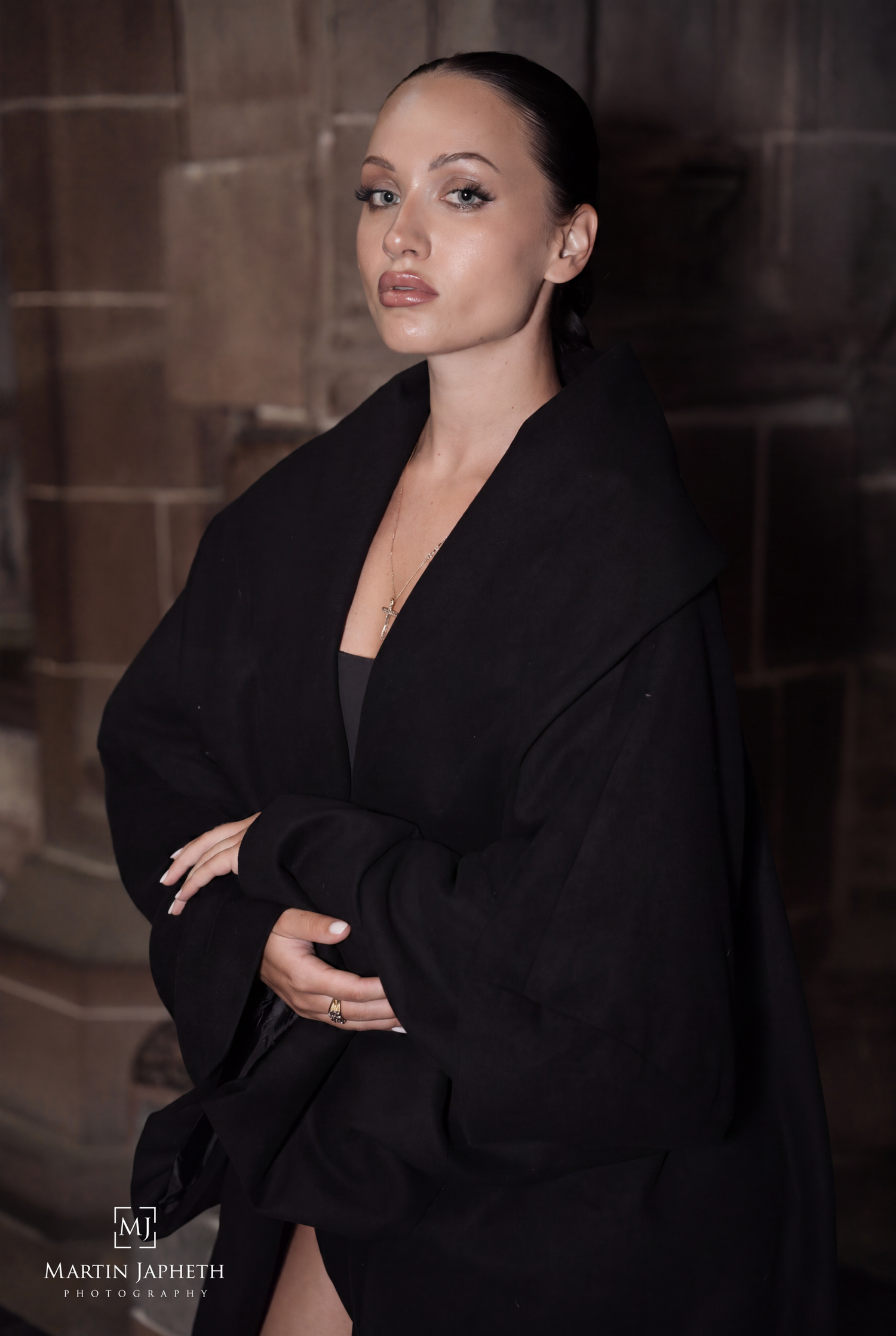 A woman in a black oversized coat poses confidently against a stone wall in a moody, dramatic portrait photo.