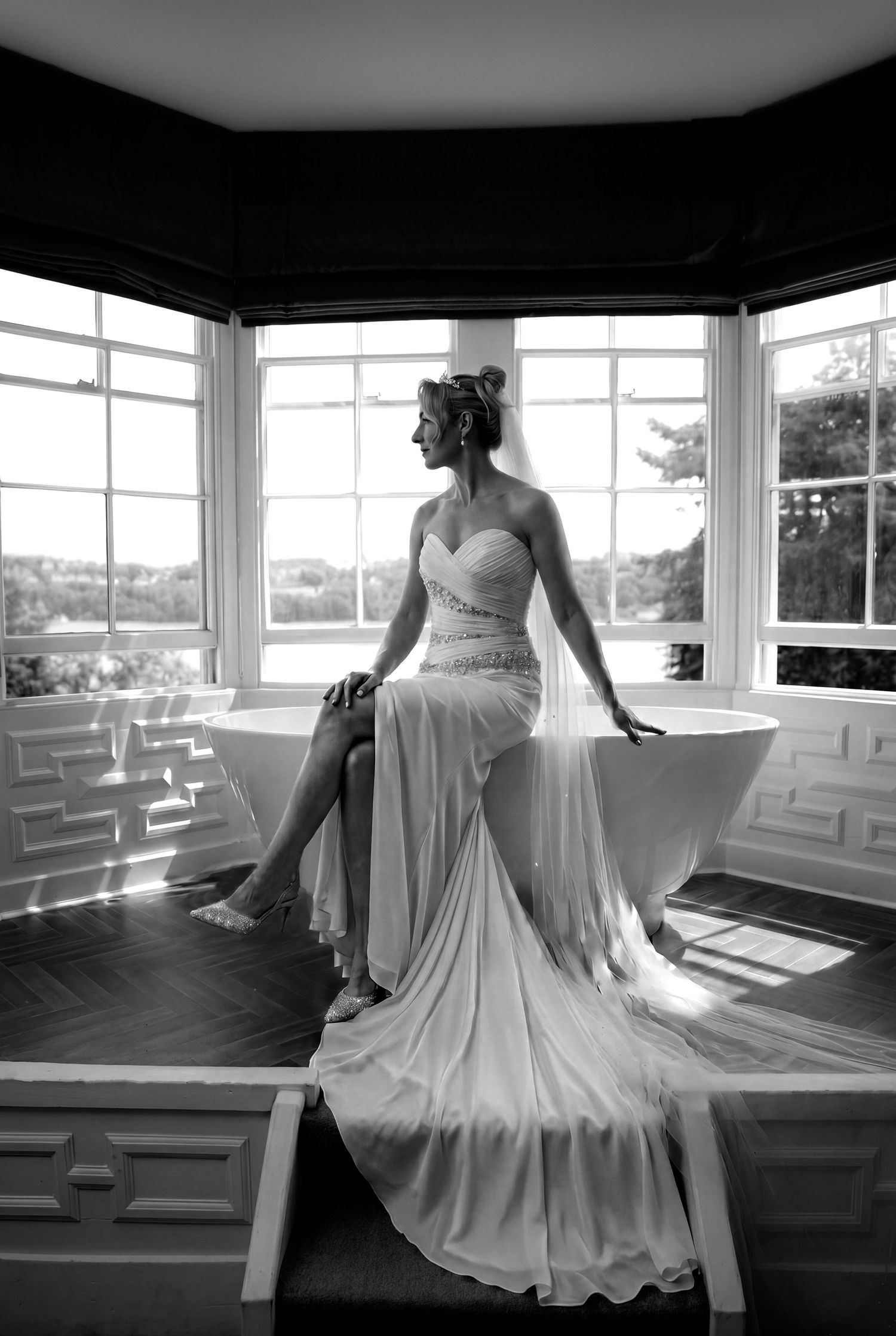 Elegant bride in white gown sitting by bay windows in black and white wedding portrait photo. Chateau Rhianfa Weddings