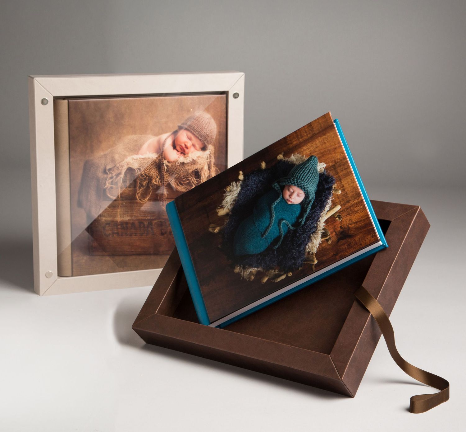 Elegant newborn photo album with teal spine displayed in a brown keepsake box, alongside a framed print of a sleeping baby.