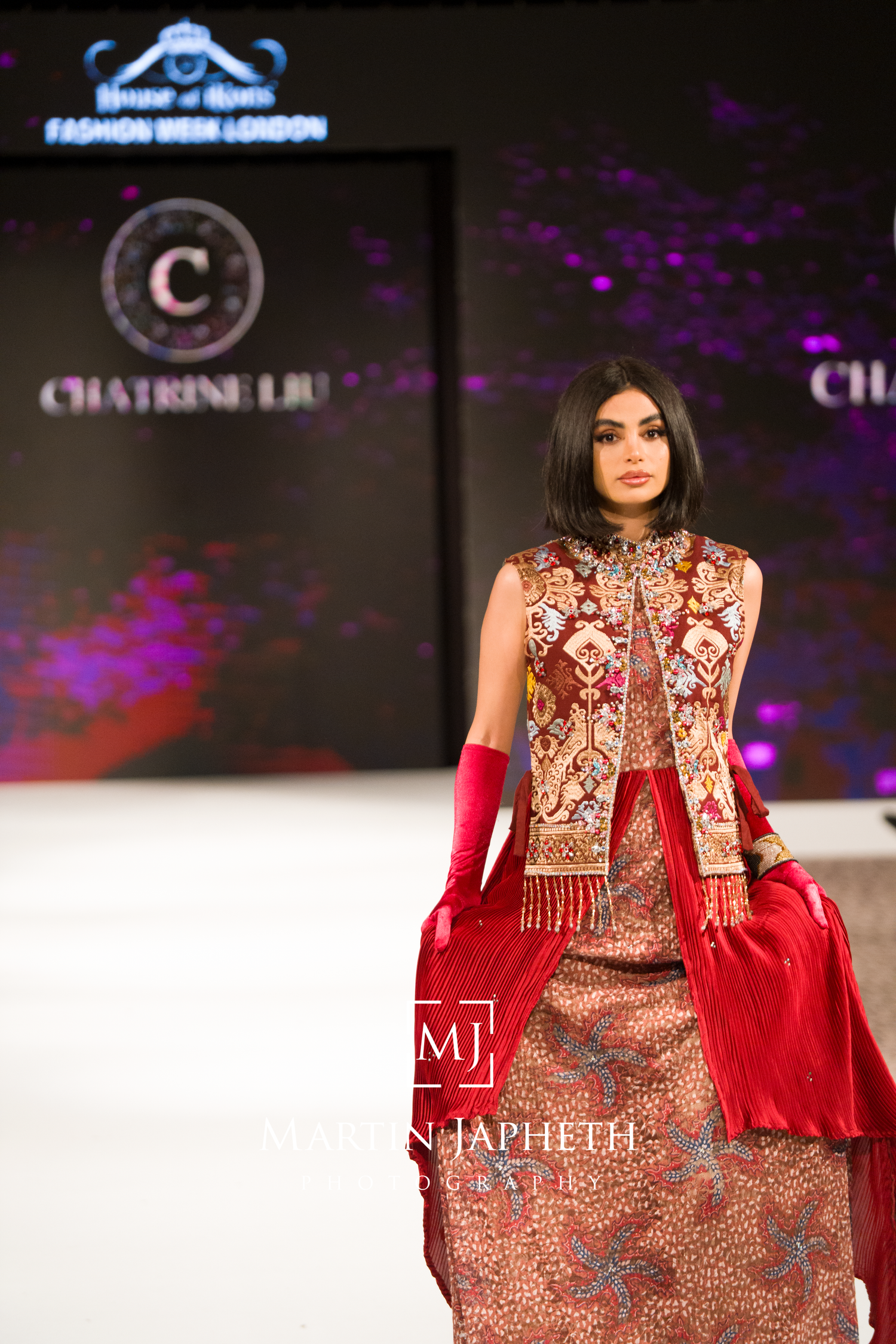 Model walks runway in ornate red and gold embroidered traditional dress with matching vest and red accessories at fashion show.