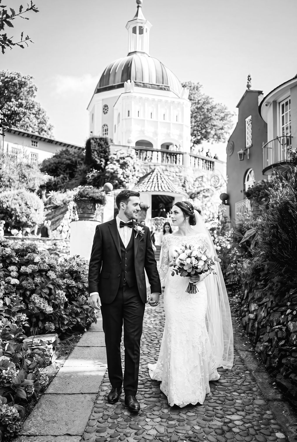 Black and white photo of a bride and groom walking hand in hand through a lush garden with a domed building backdrop.