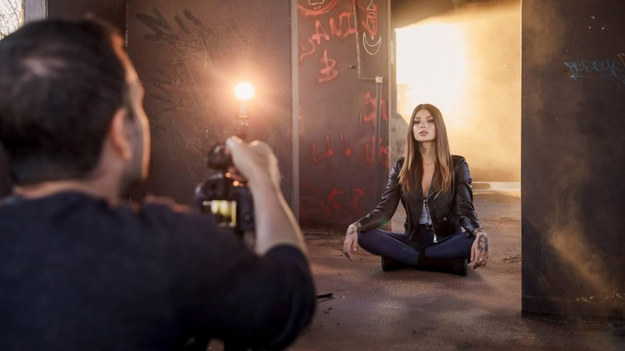 Photographer shooting model in cross-legged pose against illuminated backdrop.