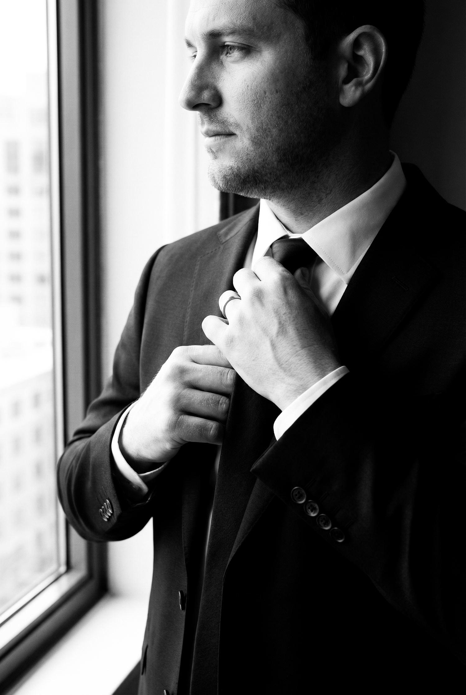 Black and white photo of a man in a suit adjusting his tie while standing by a window.