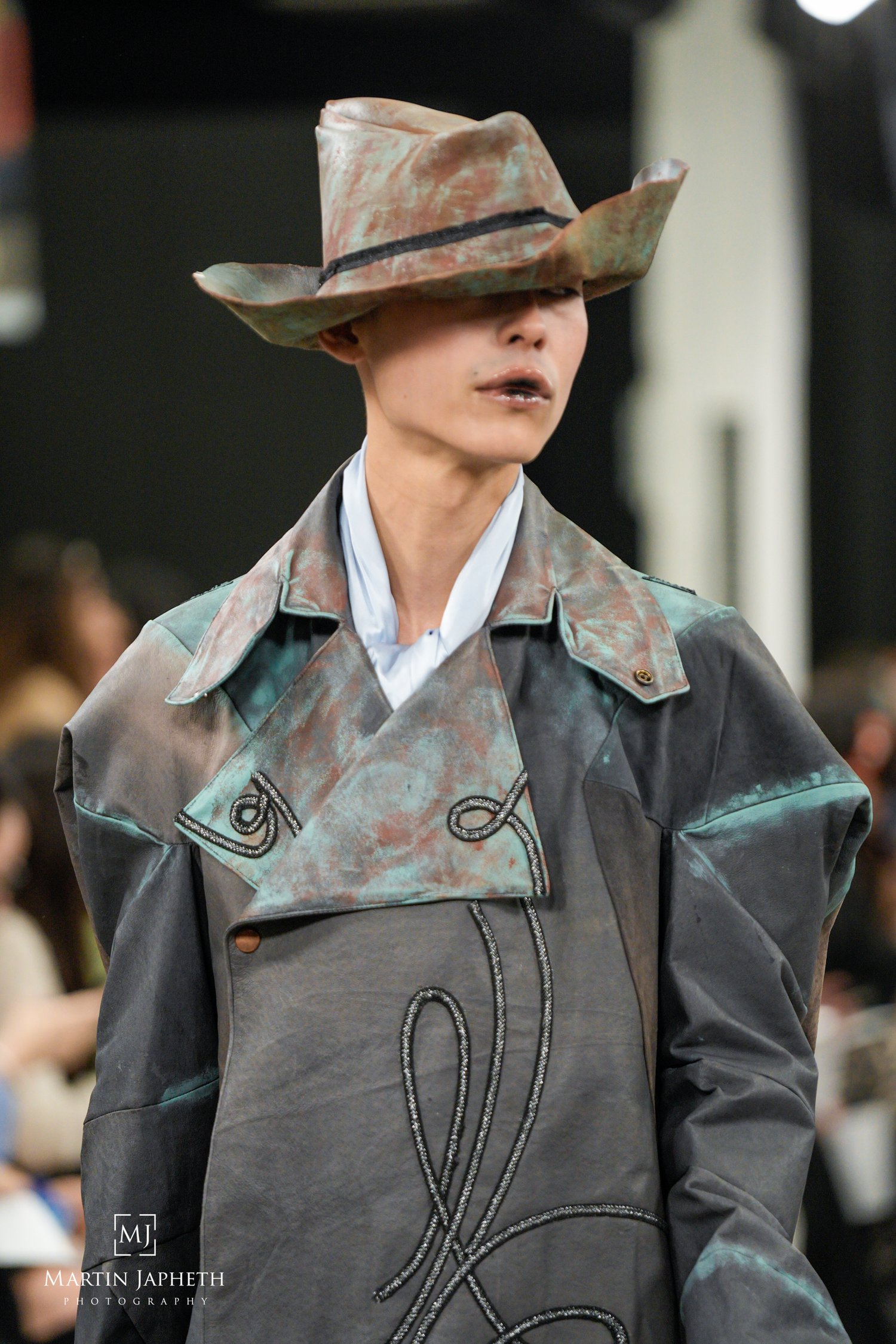 Model wearing a weathered copper-toned cowboy hat and gray coat on a fashion runway. Martin Japheth Photography