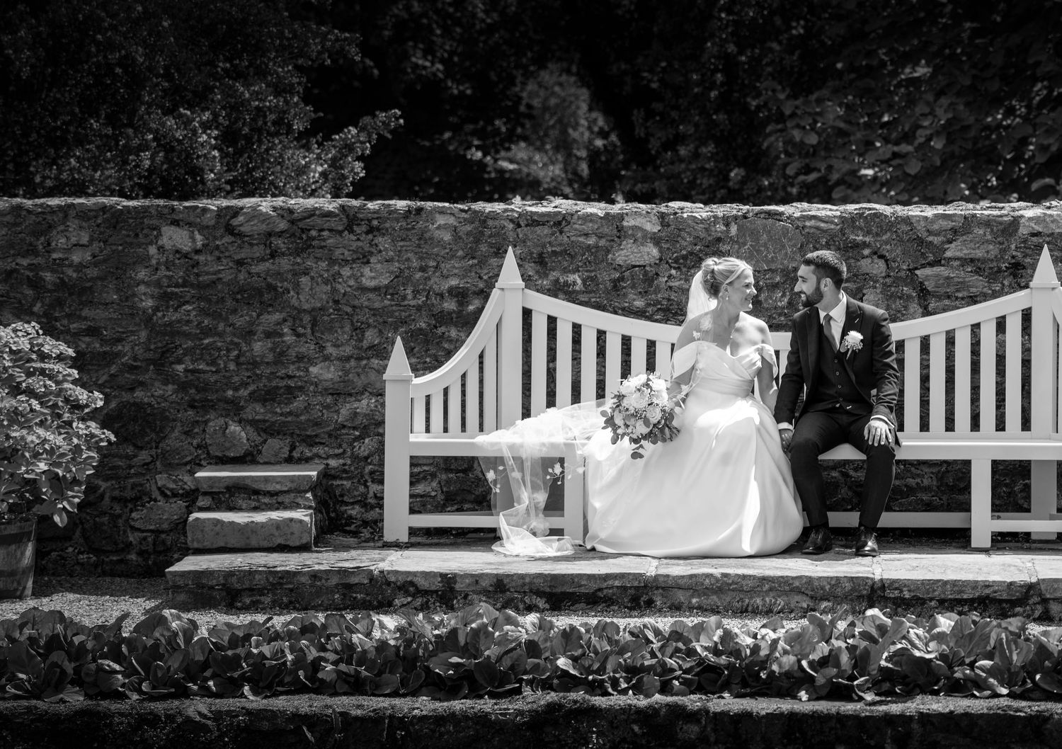 Black and white wedding photo of bride and groom sitting on a white bench in a garden setting.
