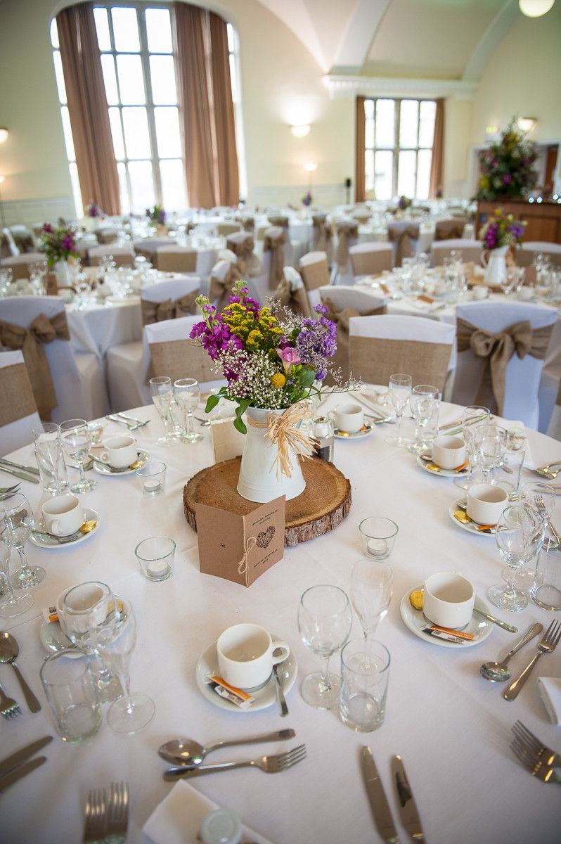 Elegant wedding reception hall with round tables, white linens, burlap chair sashes, and wildflower centerpieces on rustic wood slices.