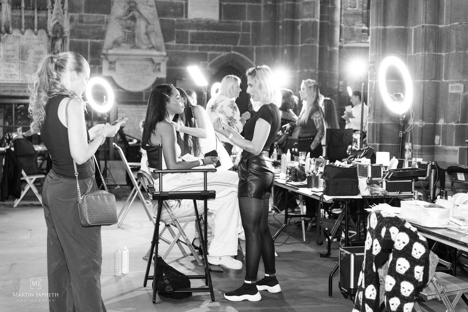 Black and white photo of models and makeup artists preparing backstage at a fashion event with ring lights.