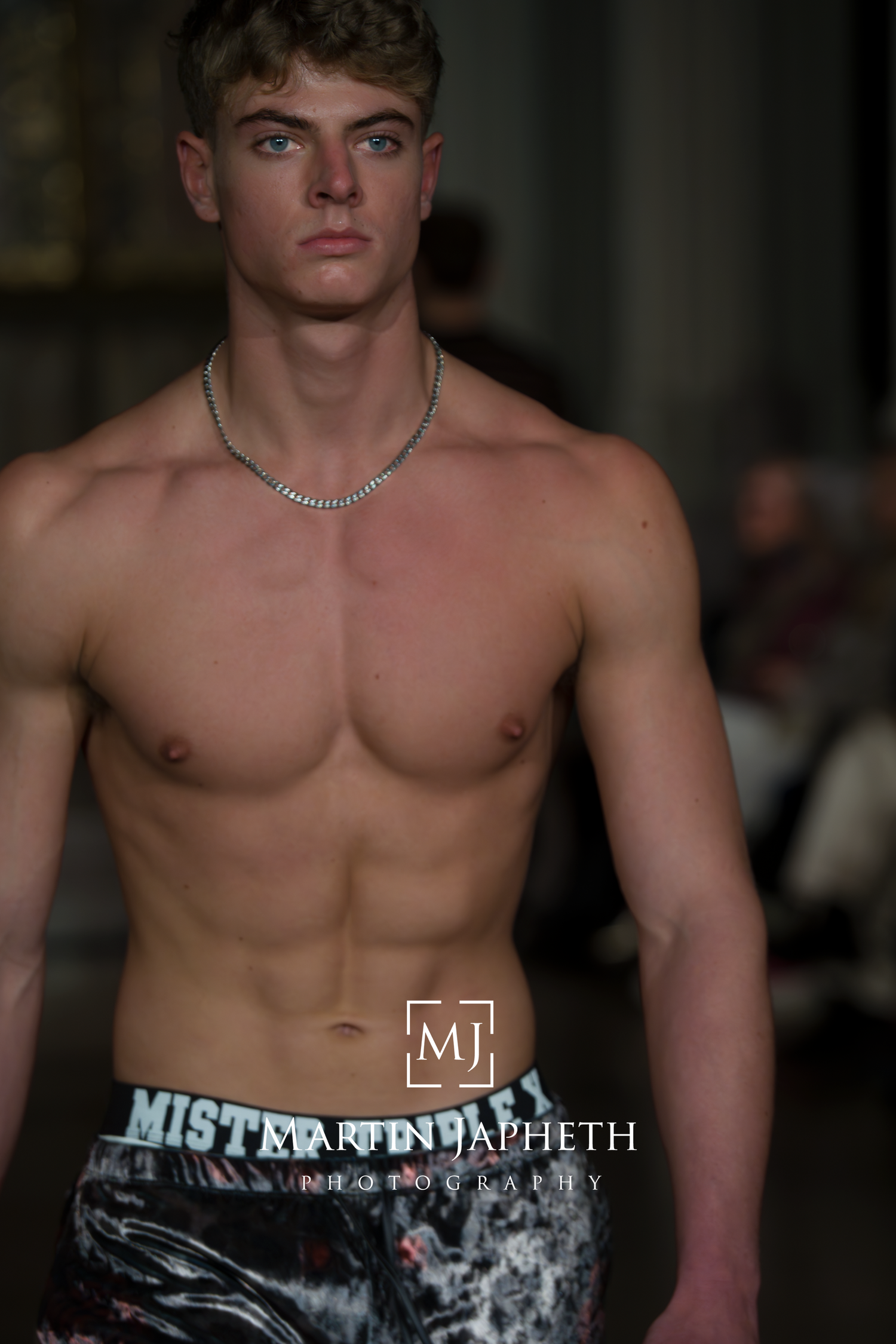 Athletic model wearing silver chain necklace and patterned Mister Jeans pants on fashion runway.