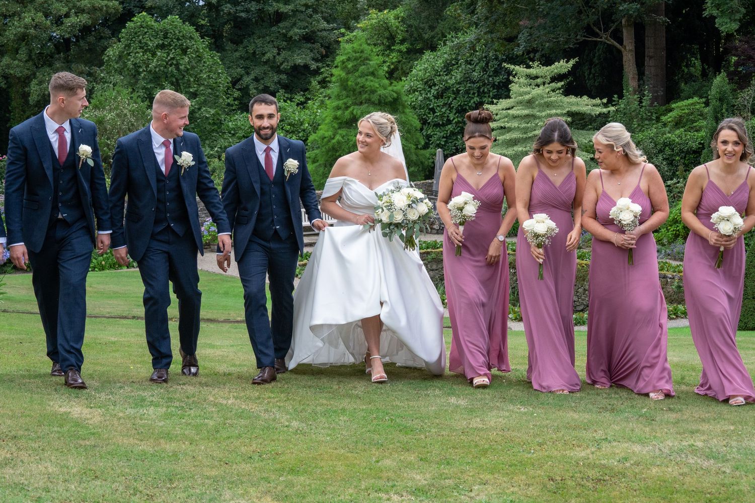 Wedding party walks outdoors, bride in white gown with groomsmen in navy suits and bridesmaids in mauve dresses.