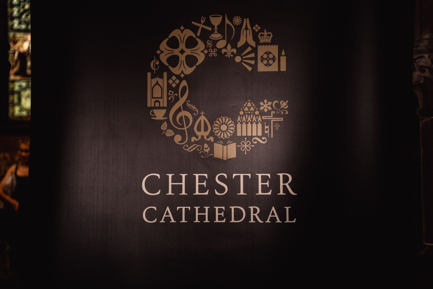 Chester Cathedral logo displayed on a dark banner with decorative golden icons arranged in a circle.