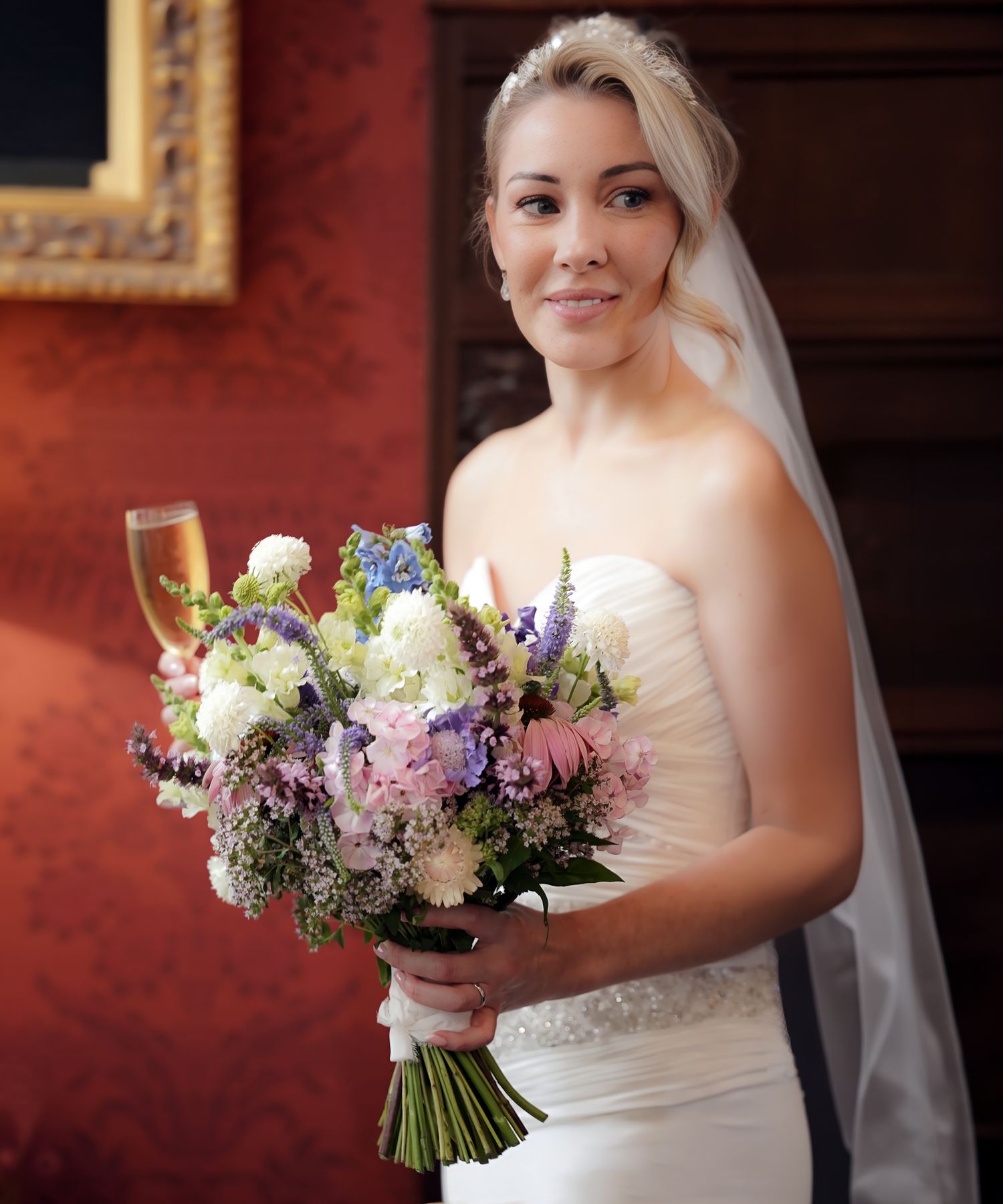 Bride in white strapless gown holding colorful bouquet of purple, pink, and white flowers with veil and champagne glass at Chateau Rhianfa