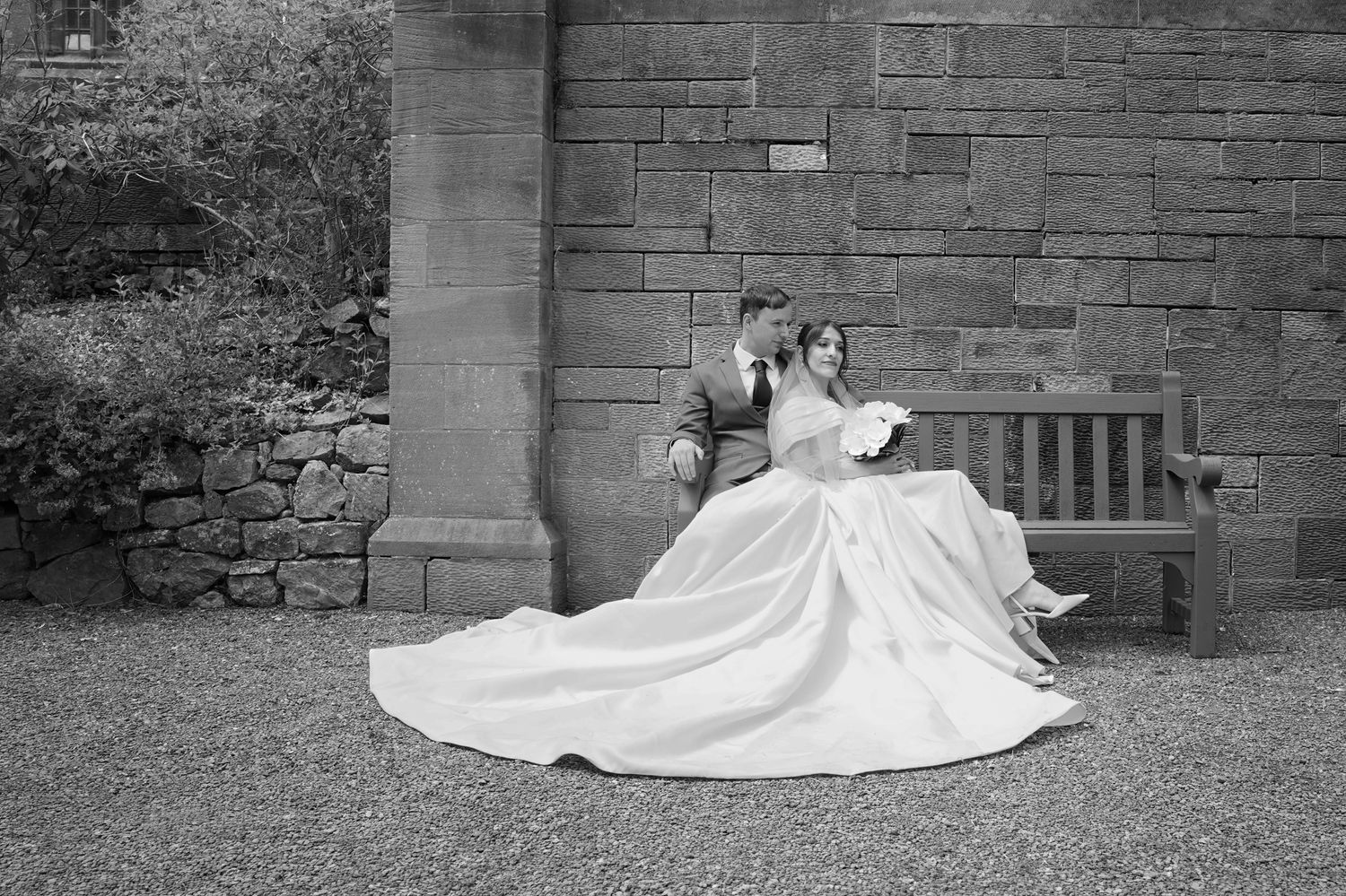 Black and white wedding photo of bride and groom sitting on a bench by stone wall in a garden.