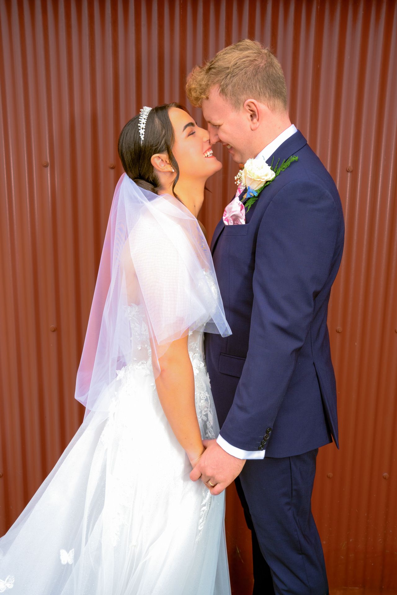 A smiling bride in a white gown and veil holds hands with a groom in a navy suit with floral boutonniere.