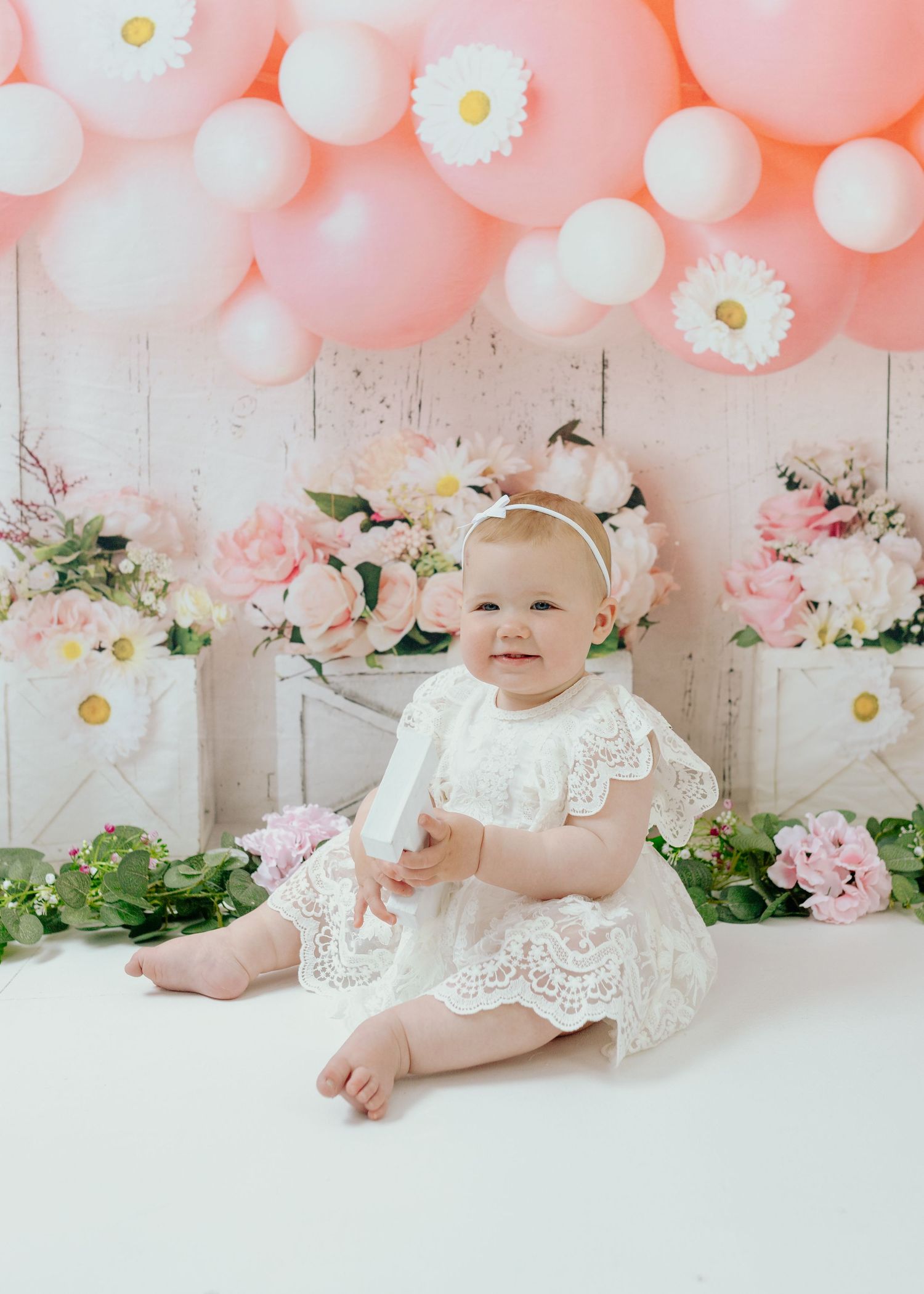 Sweet baby photoshoot setup with pink balloons, white daisies, and lace dress against rustic backdrop.