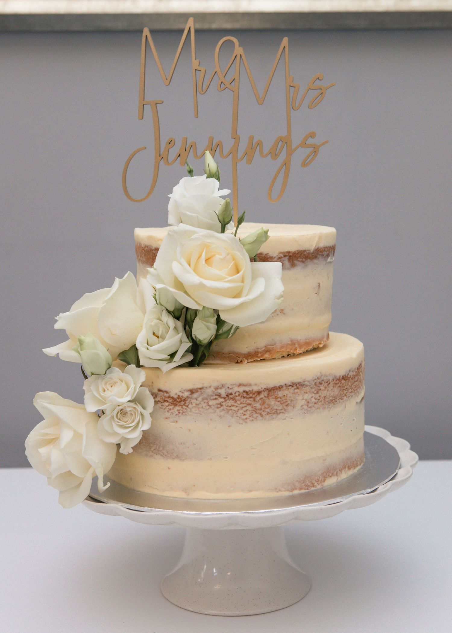Semi-naked wedding cake decorated with fresh white roses on clear cake stand.