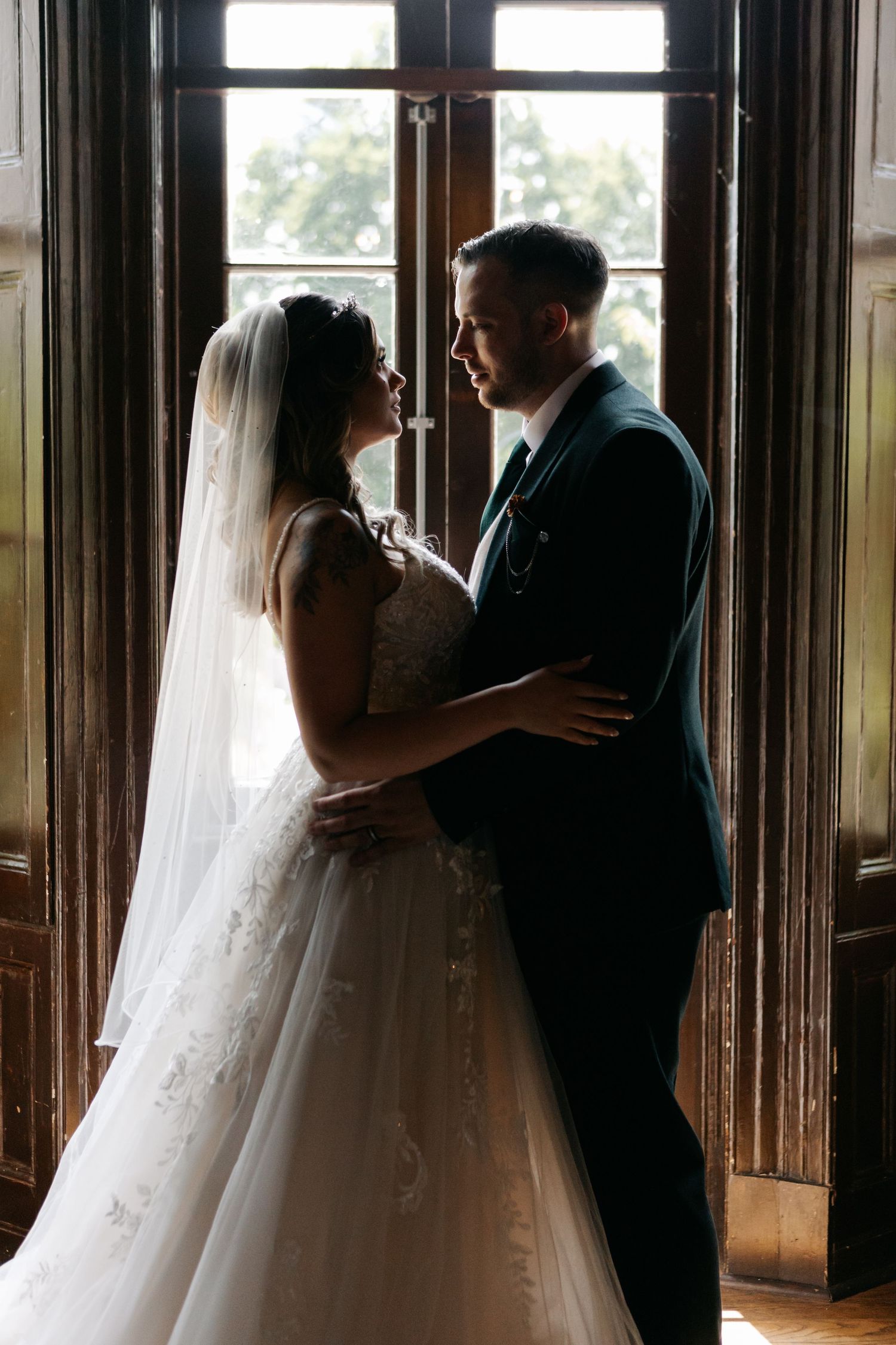 Silhouetted figures in wedding attire share an intimate moment by large windows in a wood-paneled room.