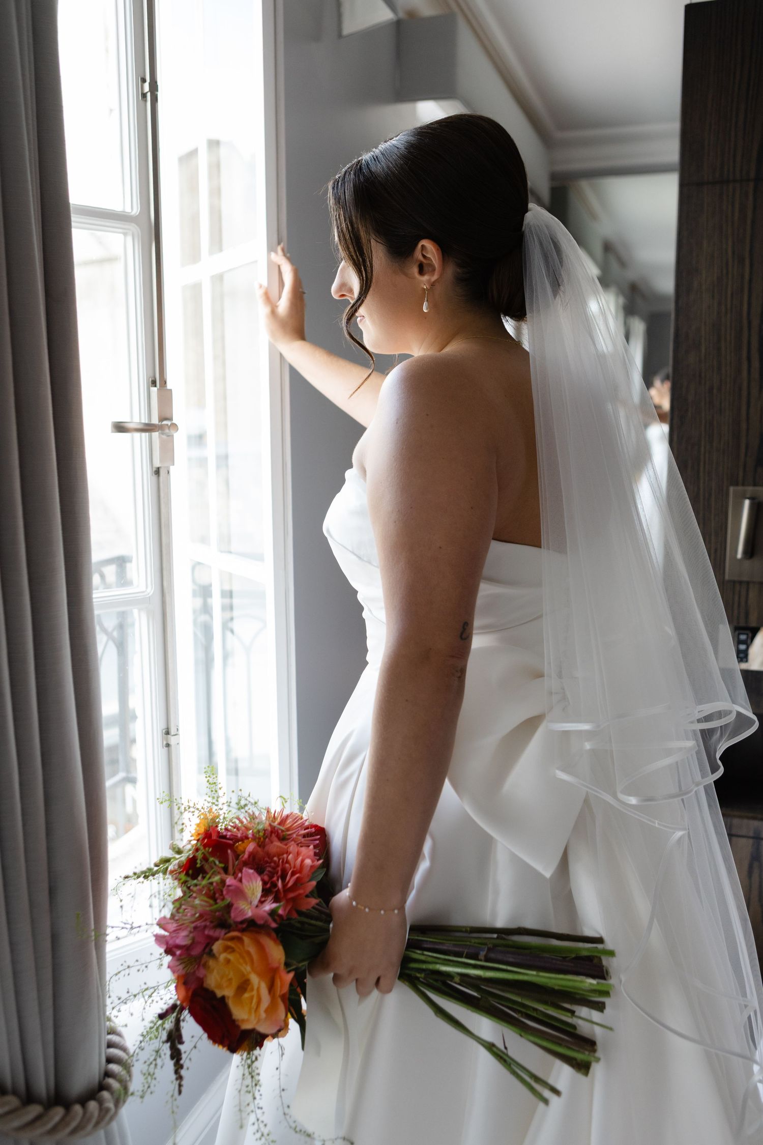 A bride in a white wedding dress holds a colorful bouquet while looking out a window.