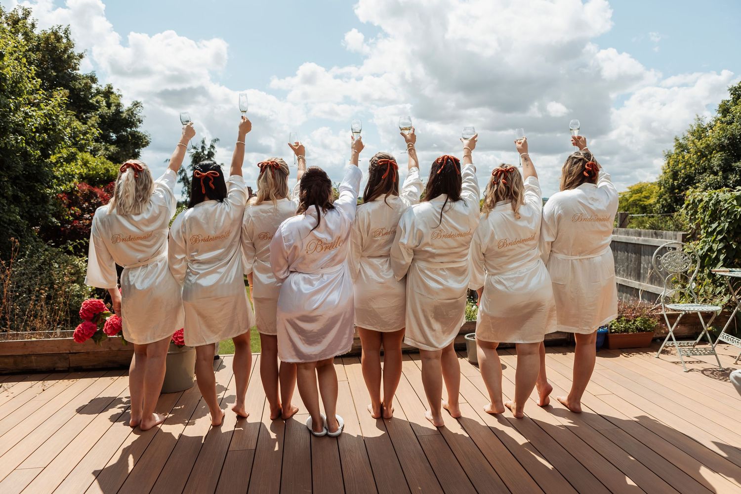 Bridesmaids in white robes pose together on a deck overlooking scenic landscape on wedding day.