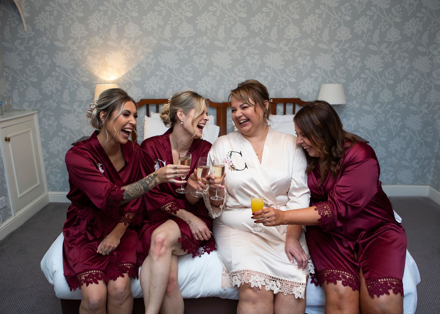 Wedding party in burgundy robes sharing drinks and laughter during preparation.