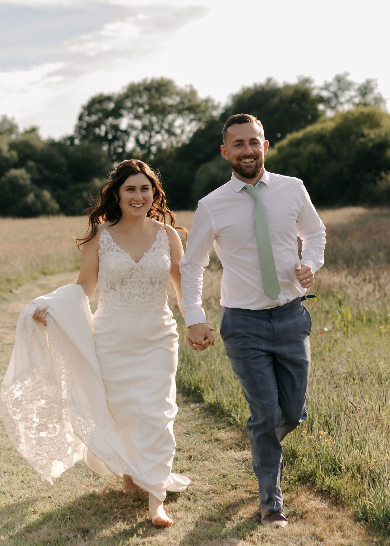 Joyful couple running through meadow with bride's dress flowing in summer breeze.
