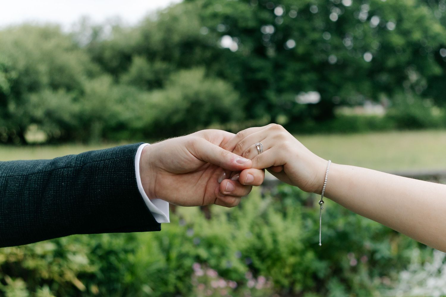 Wedding rings displayed with hands reaching across a natural outdoor backdrop.