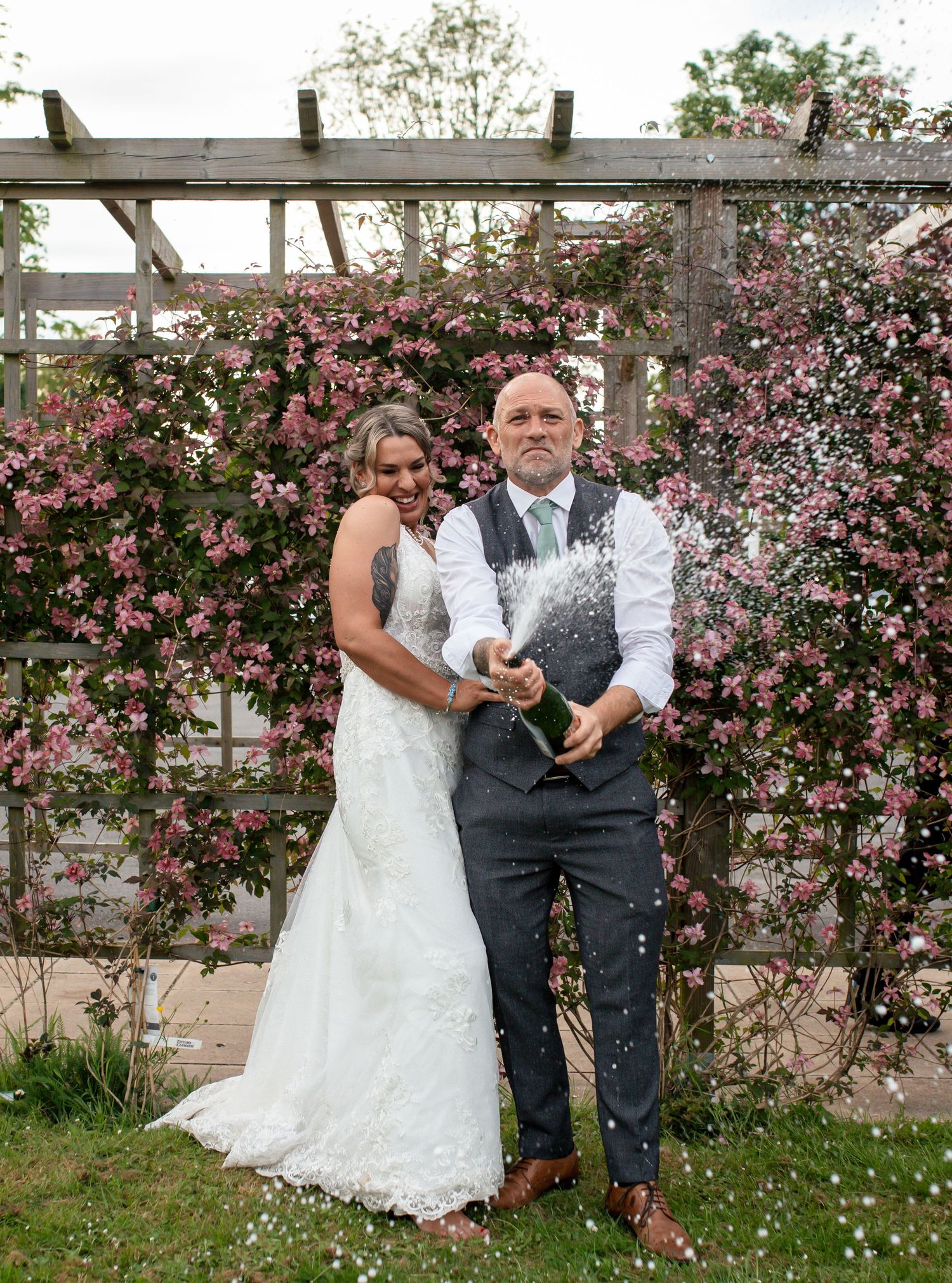 Wedding couple stands in front of blooming pink rose trellis during celebration.