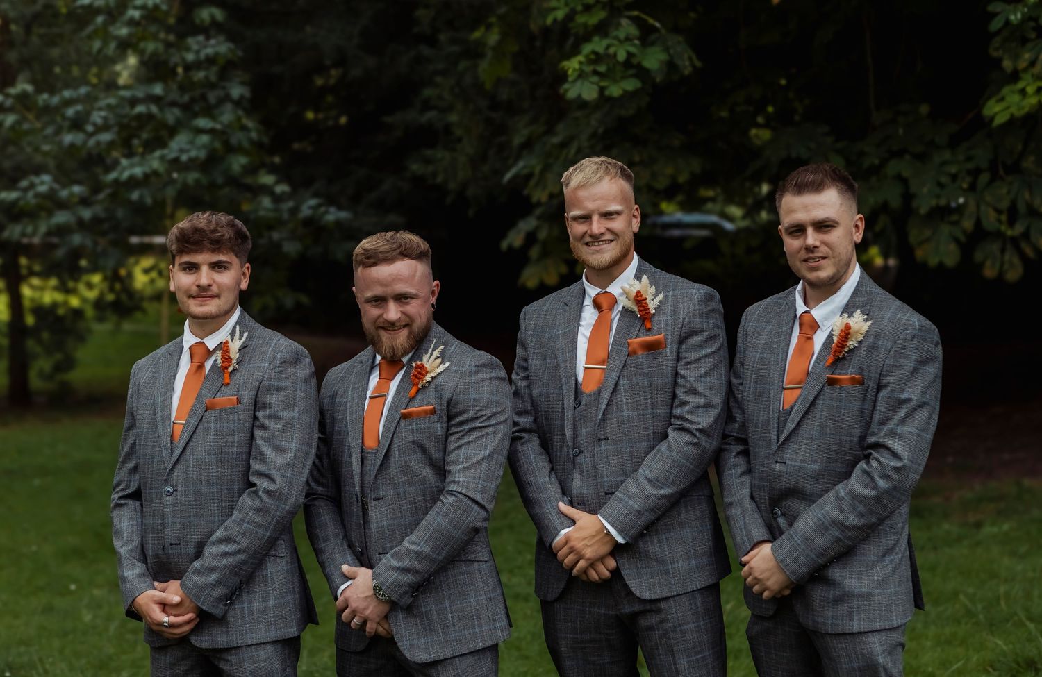 Groomsmen in matching grey suits and orange ties pose formally outdoors.