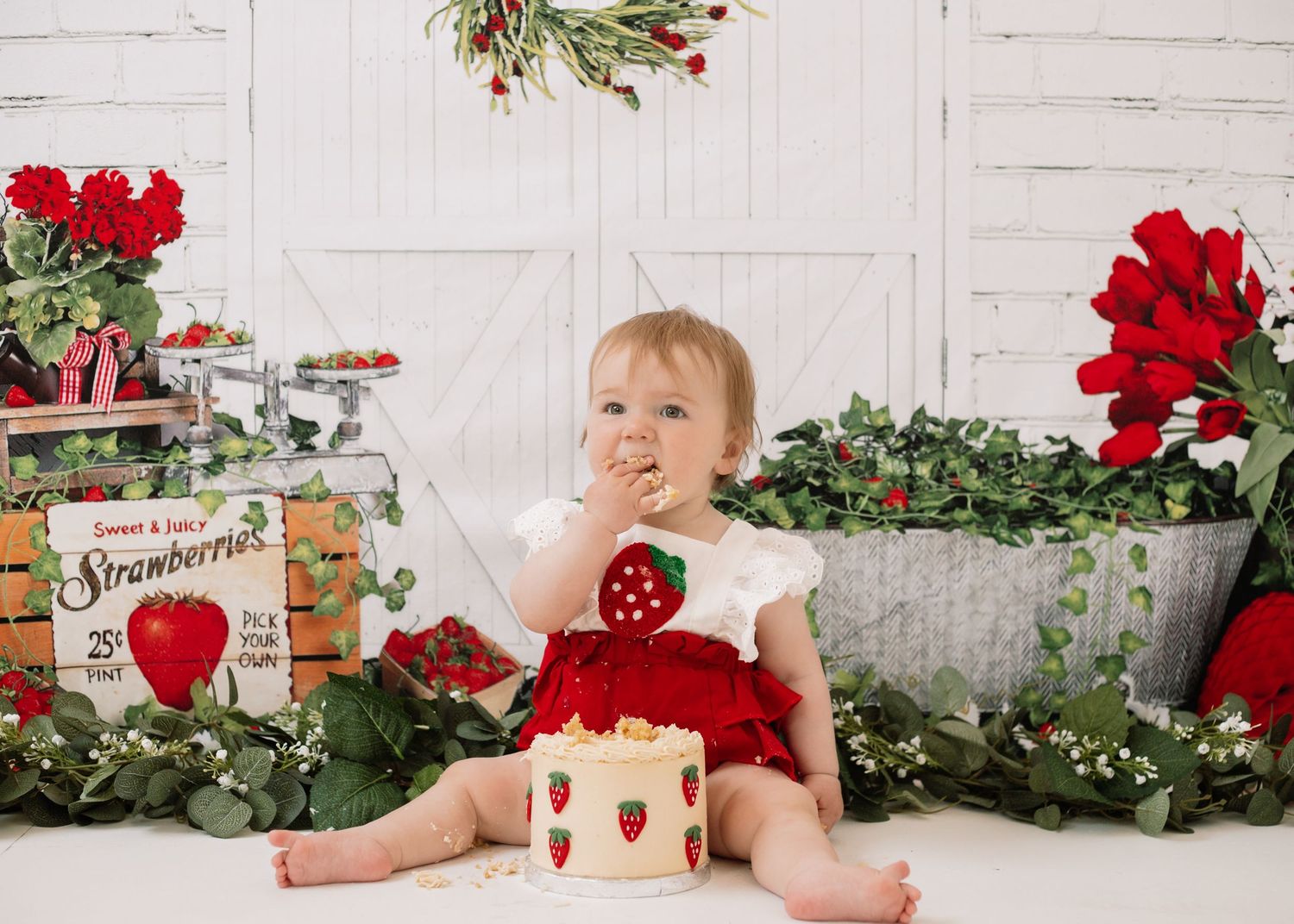 Baby in festive outfit surrounded by red flowers and strawberry themed decorations.