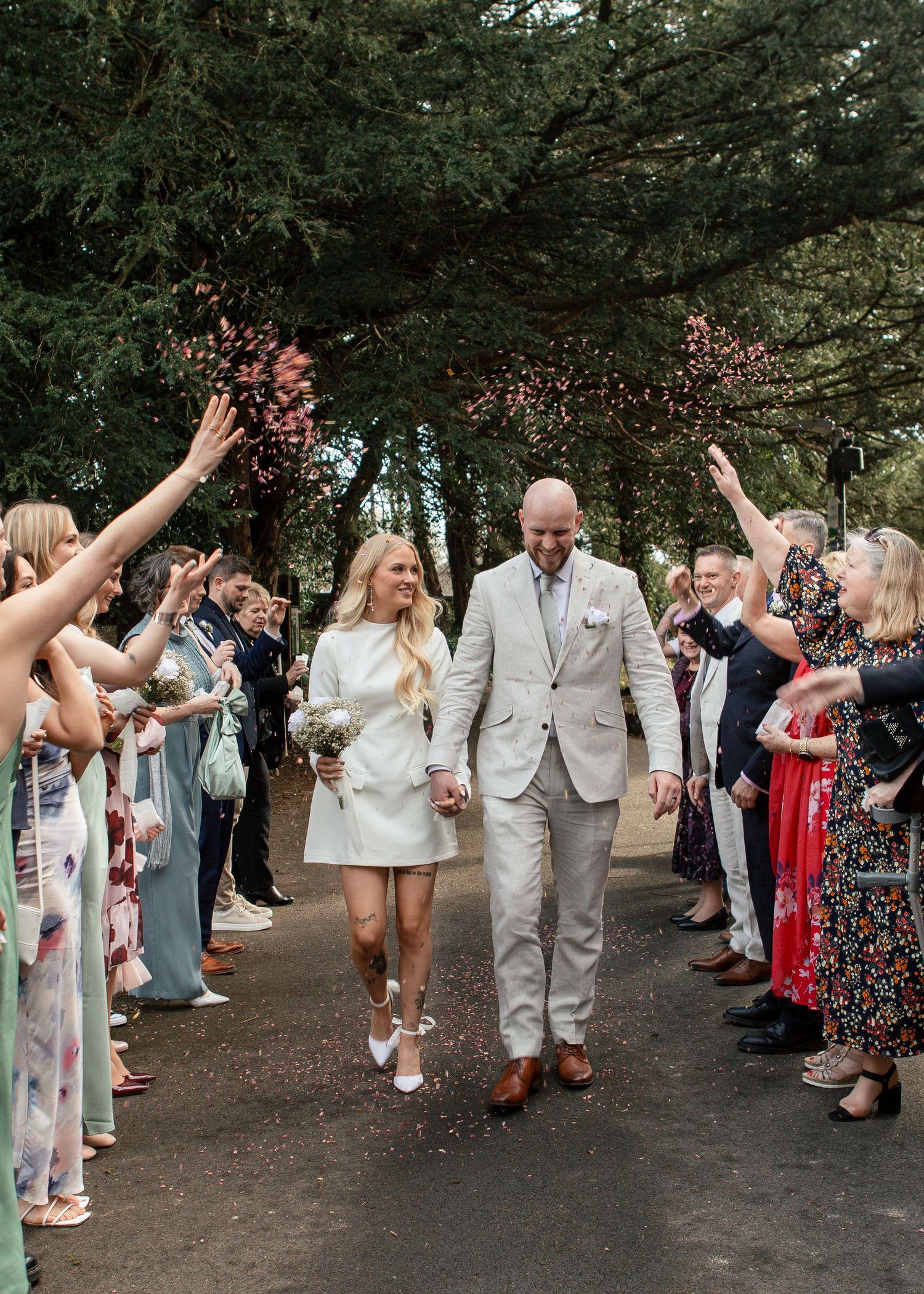 Wedding guests form a celebratory receiving line on a tree-lined path throwing confetti in the air.