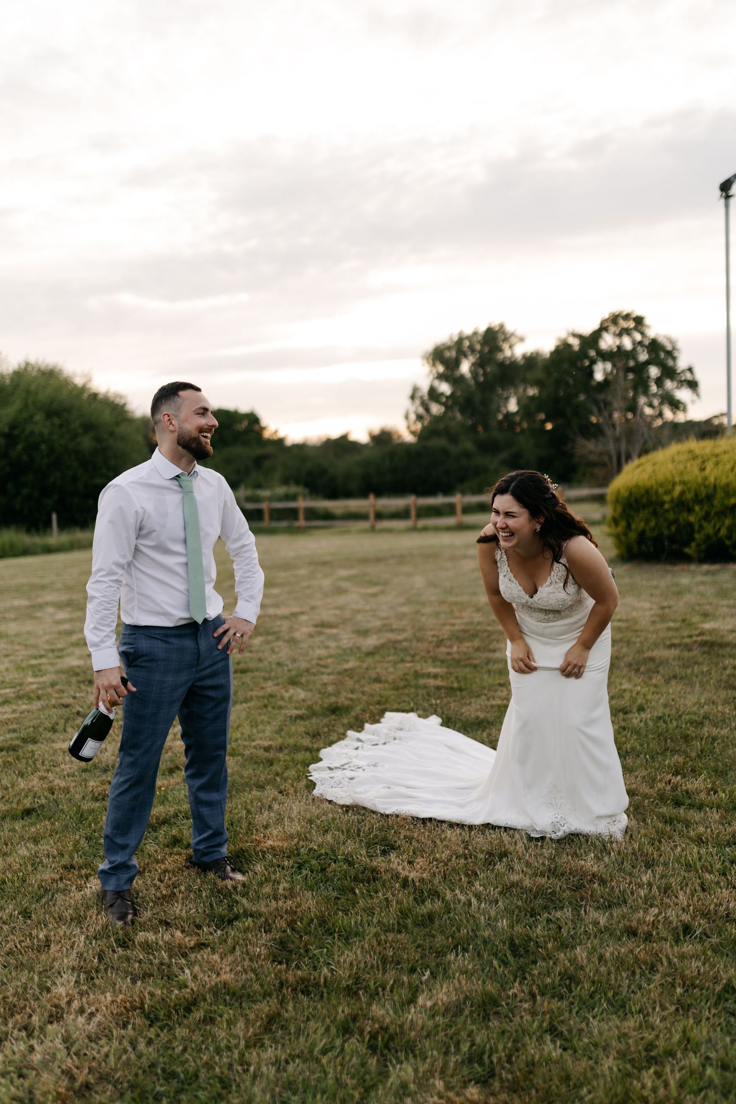 A bride in a white dress poses playfully in a grassy field at sunset.