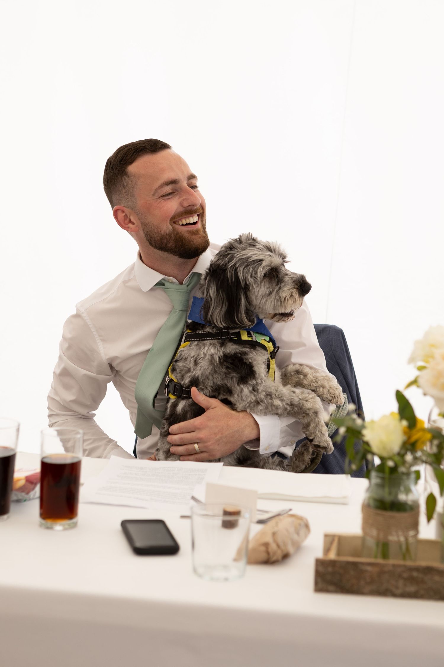 Wedding guest laughing while holding small grey dog at reception table.