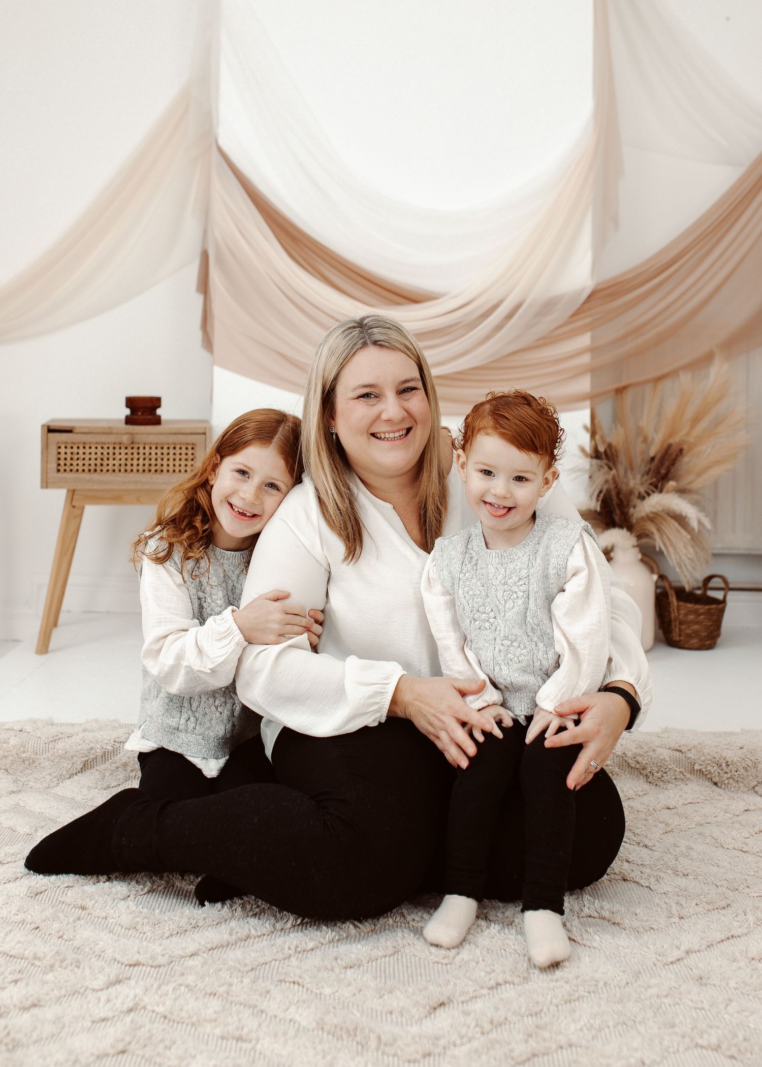 Family portrait session with matching white and gray outfits on light carpet with draped fabric backdrop.