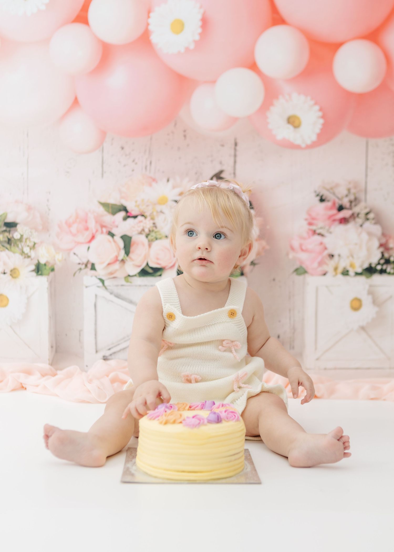 Baby in cream dress sits with yellow birthday cake against pink balloon backdrop for cake smash photoshoot.
